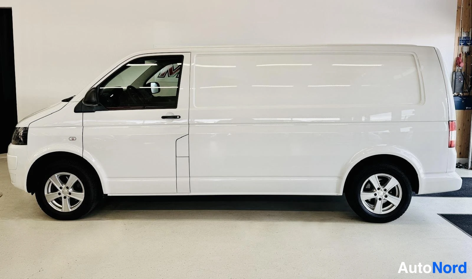 Sivulta: Valkoinen Volkswagen Transporter pakettivaunu kuvattuna sivusta vasemmalta hallissa, oven avautuva asento näkyvissä, avoin ovi paljastaa musta sisustus.