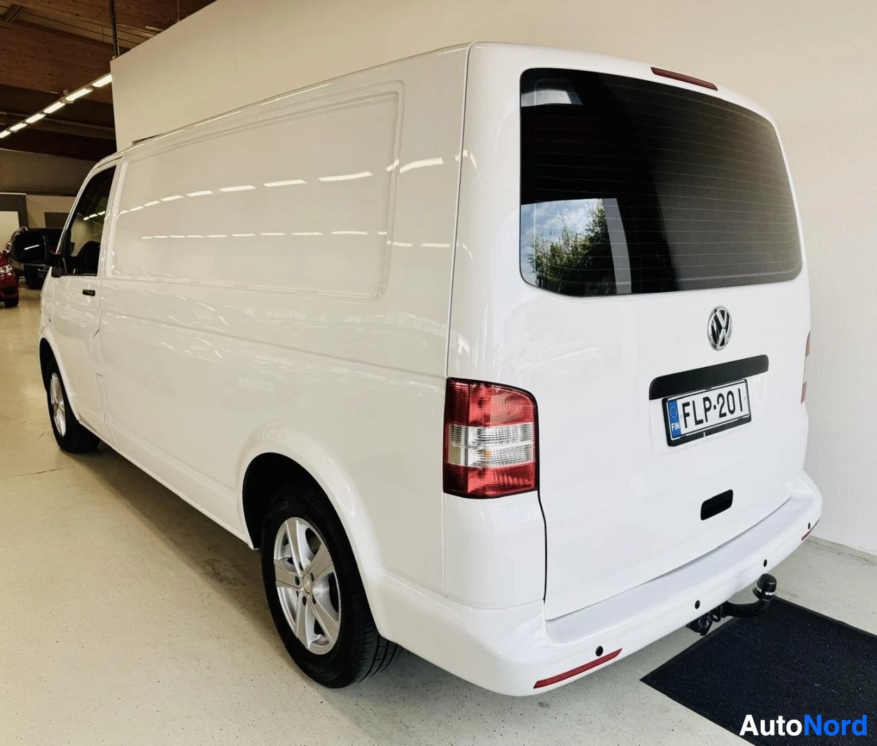 Takaviisto: Valkoinen Volkswagen Transporter pakettivaunu kuvattuna takakolmesta oikealla hallissa, takaisinpäin näkymä, rekisterikilpi ja takavalo näkyvissä.
