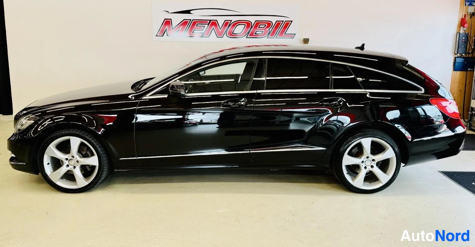 Sivulta: Musta Mercedes-Benz CLS Shooting Brake sivuprofiili oikealta, valkoiset vanteet, pitkä takalohko, hallissa Menobil-logon kanssa