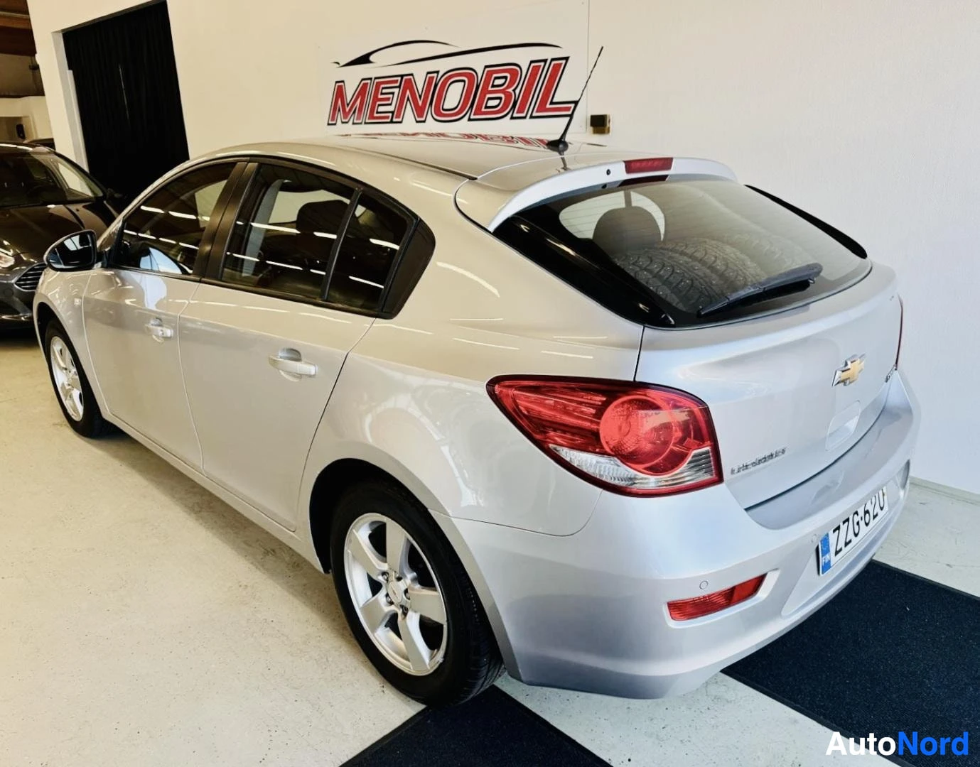 Takaviisto: Valkoinen Chevrolet Cruze, takaosasta nähtynä oikealle osoittavasta kulmasta. Vasen ovi auki, rekisterikilpi FG06BZO näkyvissä, punaiset takavalaisetimet ja val