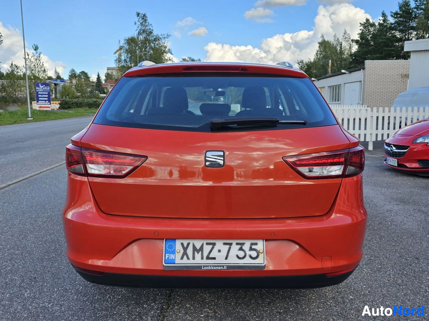 Takaa: Oranssin farmariauton takaosa, auto on kuvattu suoraan takaa. Autossa on suomalainen rekisterikilpi. Taustalla näkyy puita ja sininen taivas.