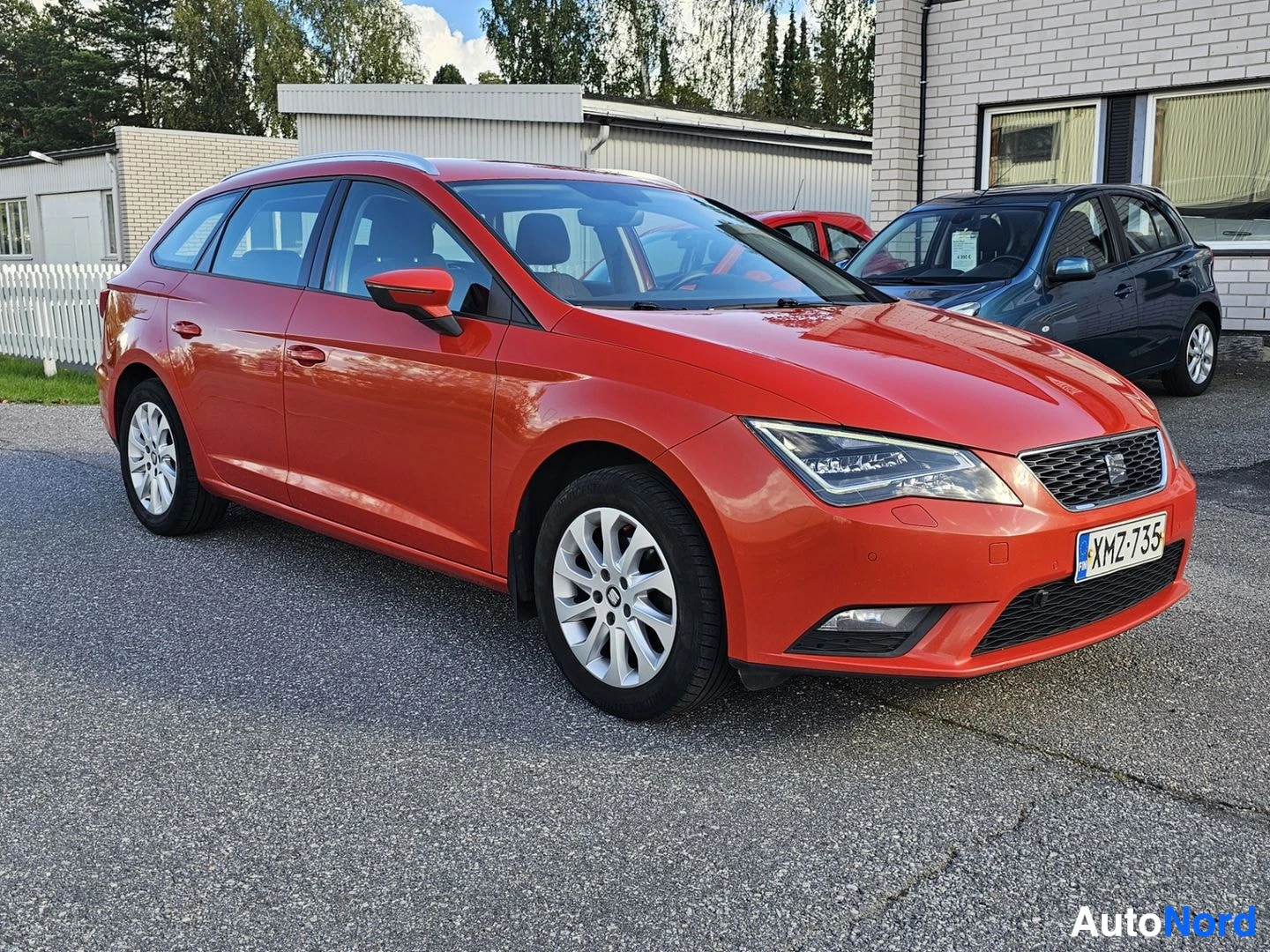 Etuviisto, nokka oikealle: Oranssi SEAT Leon -farmariauto pysäköitynä parkkipaikalle, edessä oikea kolmiokulma.