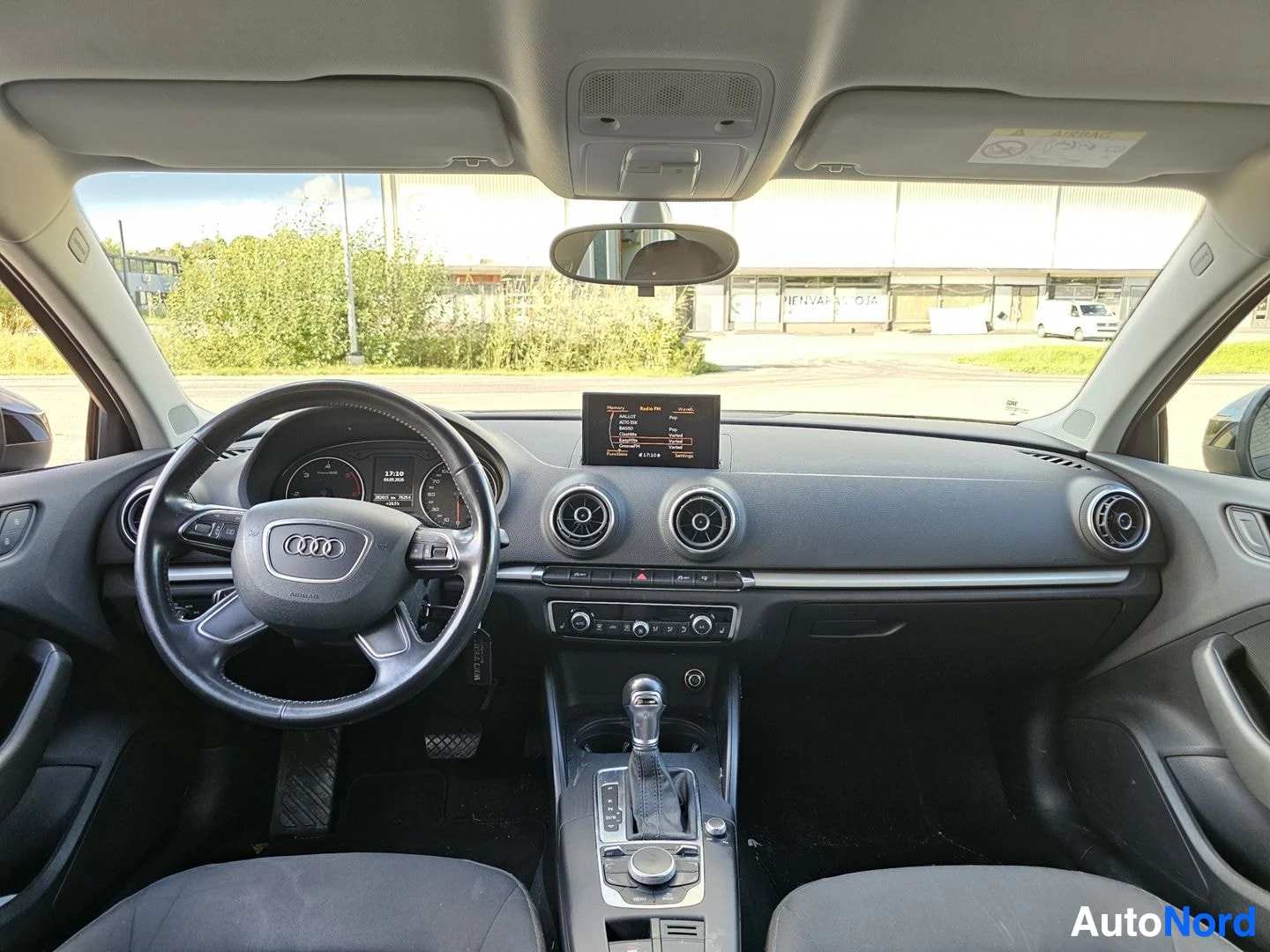 Ohjaamo: Sisätila edestä, musta kojelauta, keskellä näyttö ja ilmastoinnin säätimet, automaattivaihteiston valitsin.