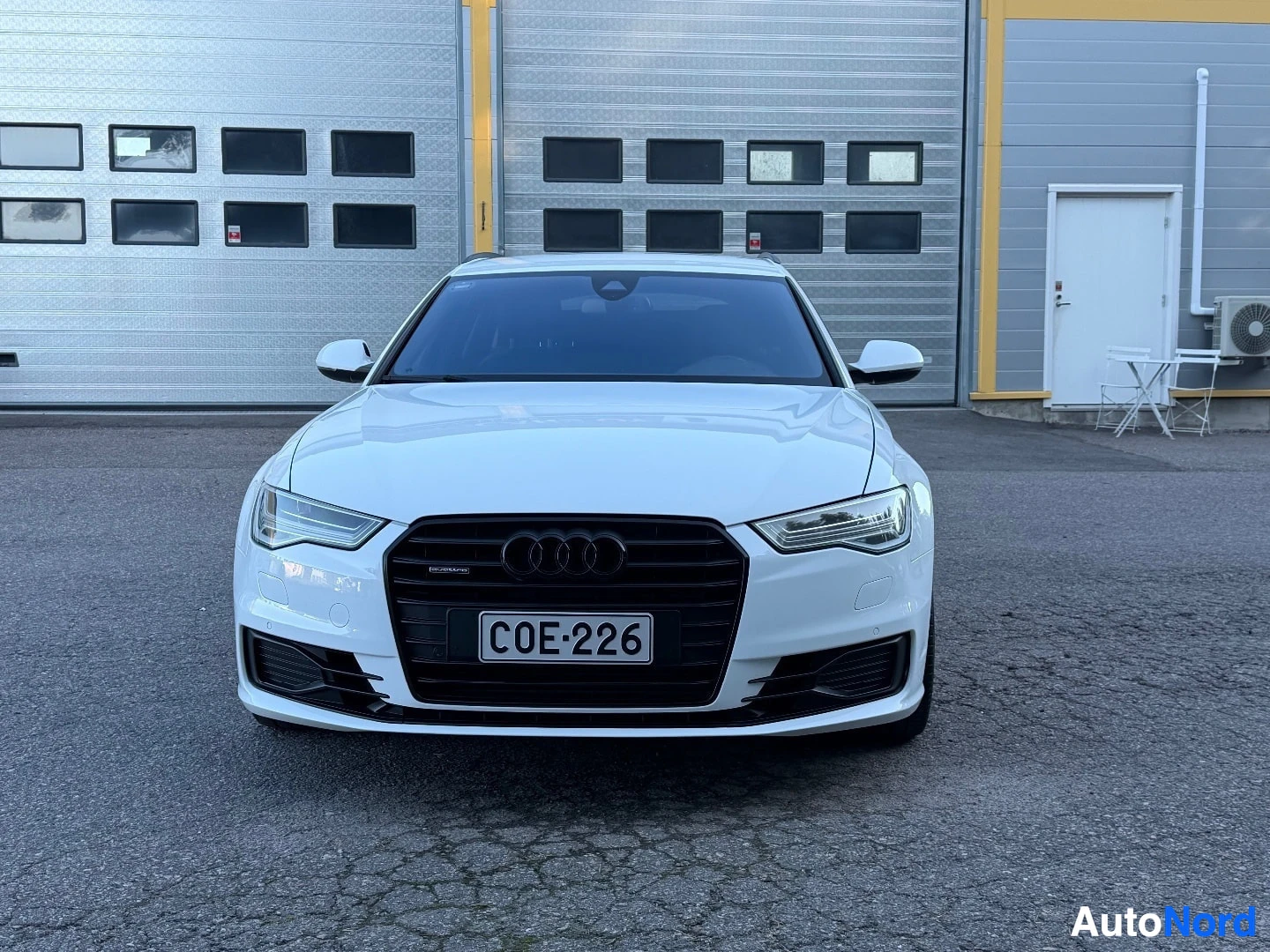 Edestä: Edestä kuvattu valkoinen Audi henkilöauto, jossa on tumma etusäleikkö ja rekisterikilpi. Auto on pysäköity harmaalle asfaltille suuren harmaan autotallin edessä