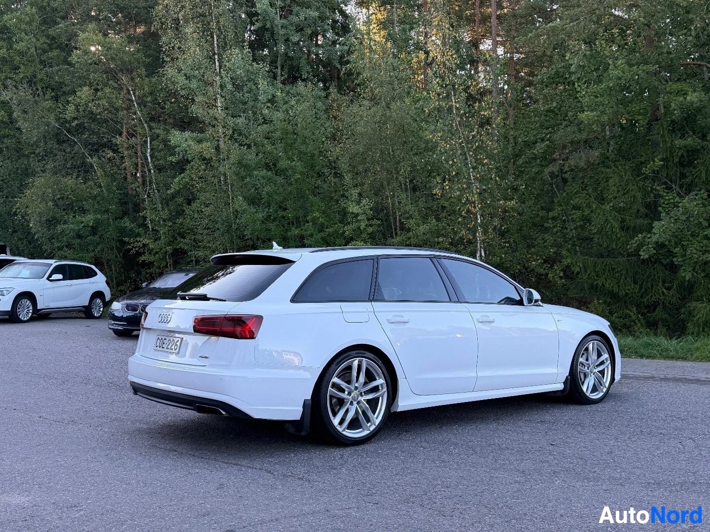 Takaviisto: Valkoinen farmariauto, jossa on rekisterikilpi "", on pysäköity ulkona puurivin edustalle. Ajoneuvossa on monipuolaiset kevytmetallivanteet.