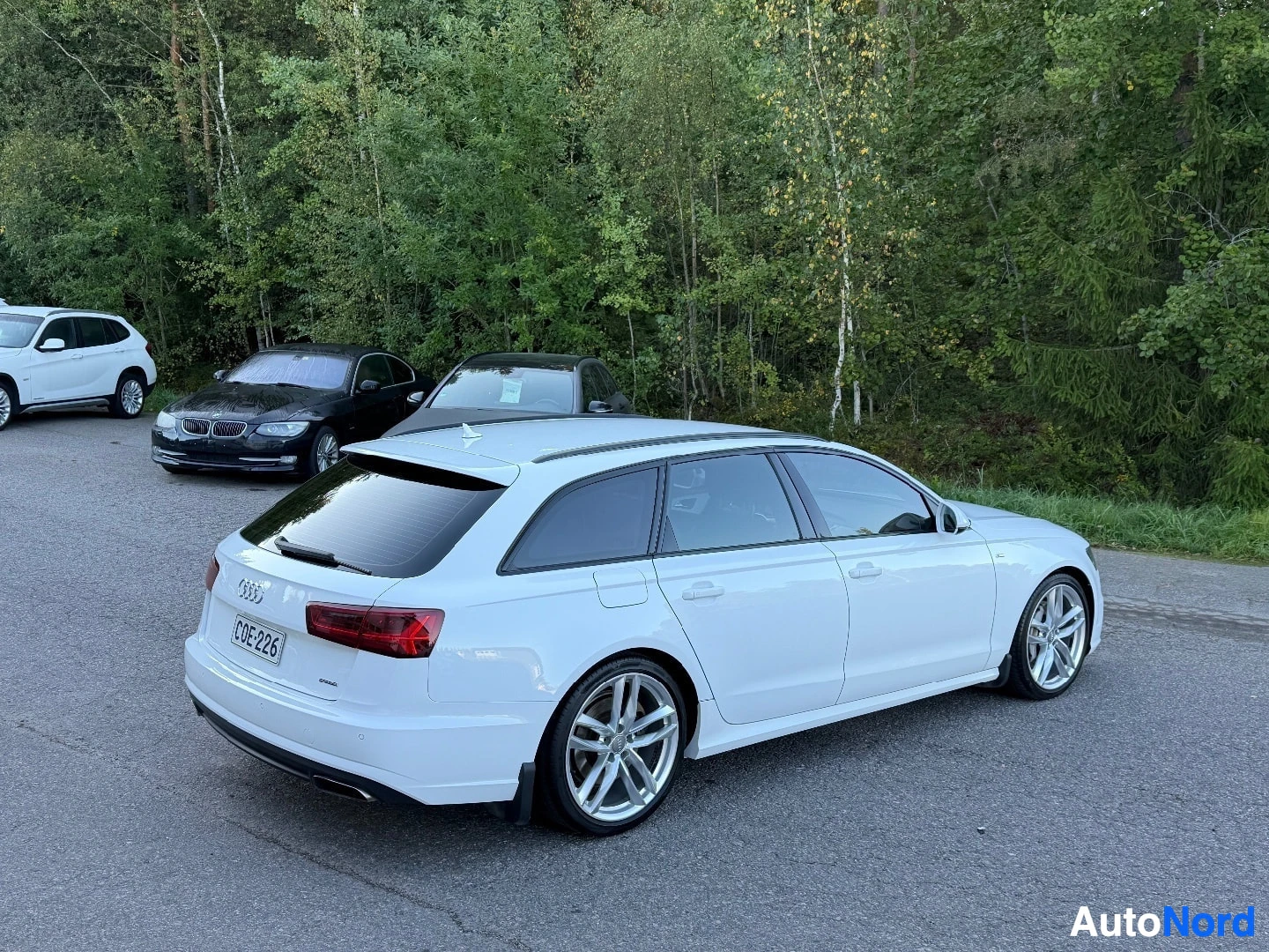 Takaviisto: Valkoinen Audi farmari, rekisterikilpi, pysäköitynä tien poskessa metsäisen taustan edessä. Auton takaosa ja oikea kylki näkyvät, vasen kylki ei. Auto on käänne