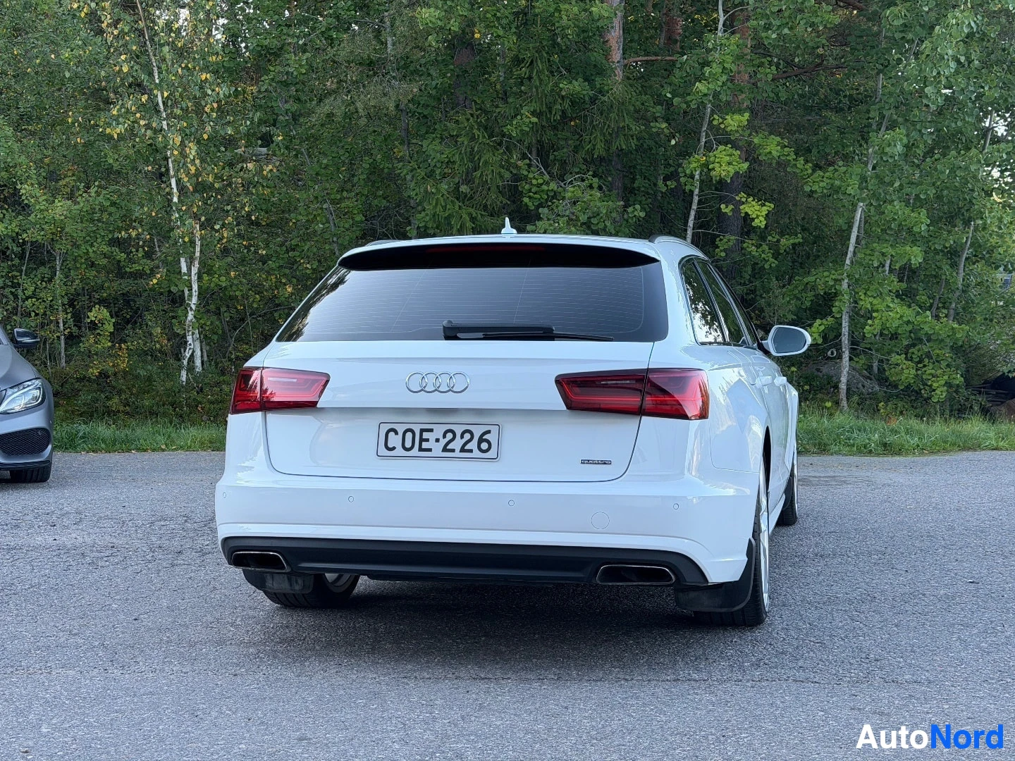 Takaa: Valkoinen Audi farmari, rekisterikilpi, takaa kuvattuna. Kaksi pakoputken ulostuloa näkyvissä. Auton vasemmalla puolella on osittain näkyvissä tumma auto.