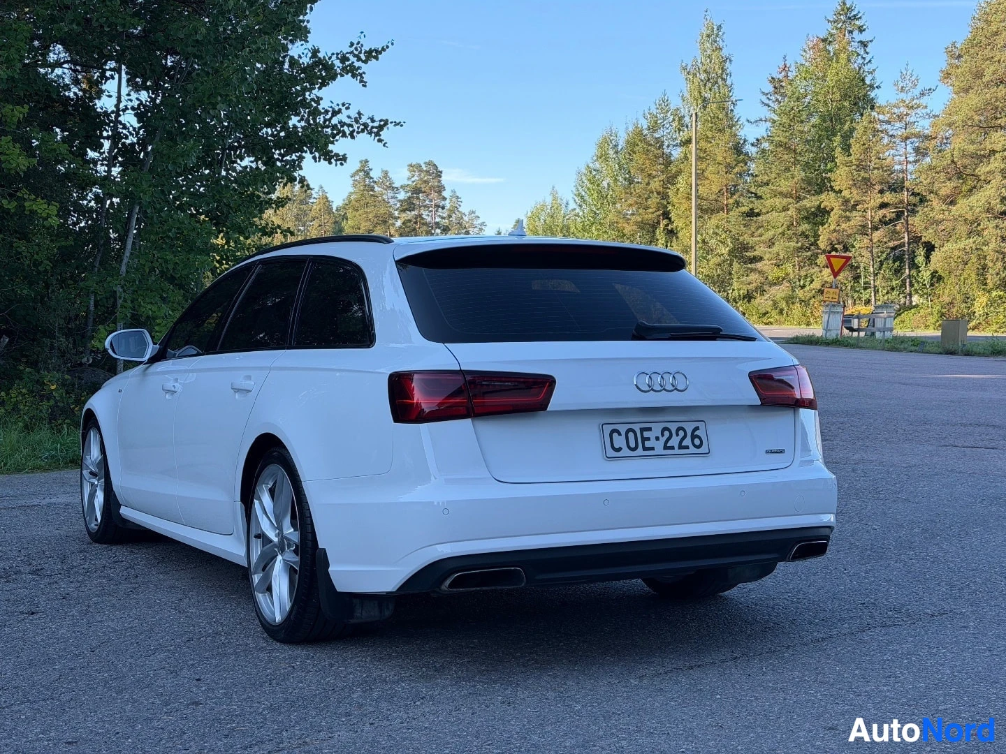 Takaviisto: Valkoinen Audi farmari, rekisterikilpi, pysäköitynä tien poskessa metsäisen taustan edessä. Auton takaosa ja vasen kylki näkyvät. Auto on käännetty vasemmalle.