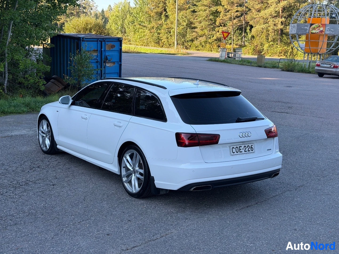 Takaviisto: Valkoinen Audi farmari, rekisterikilpi, pysäköitynä tien poskessa. Auton takaosa ja vasen kylki näkyvät. Taustalla on puita, sininen kontti ja tienvarsiopasteit