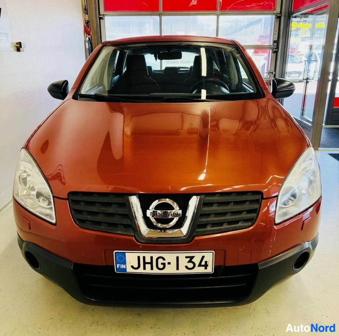 Edestä: Oranssi Nissan Qashqai näkyy edestä, hallissa. Näkyy etulasit, vetokoukku, etuvalot ja Nissan tunnus etusäleikössä.