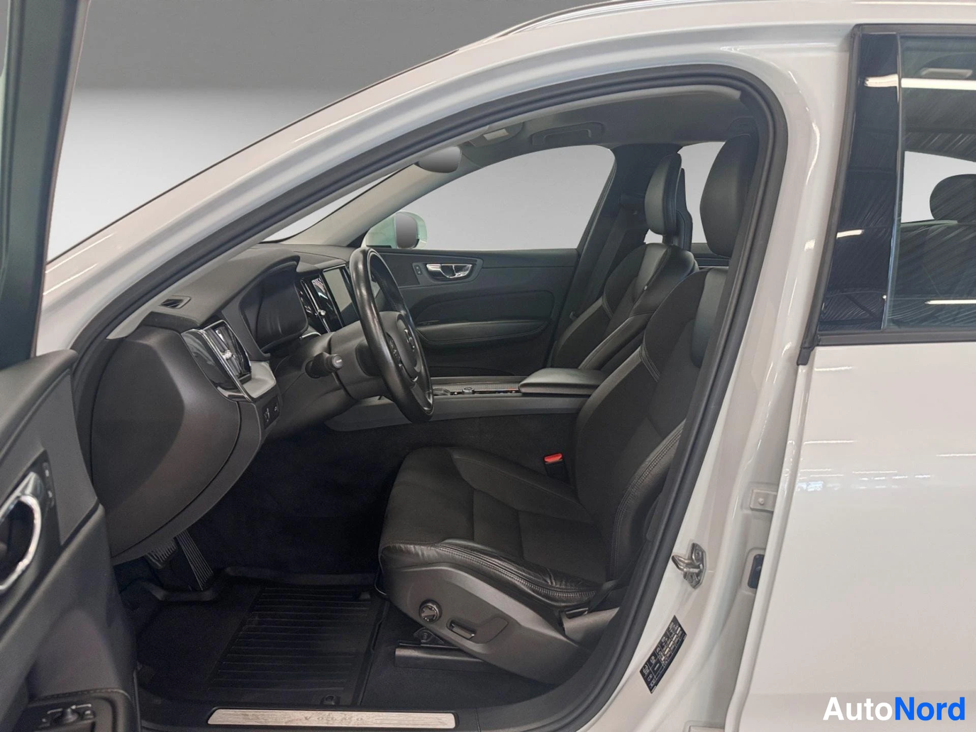 Etuistuimet: Volvo XC60:n avoin kuljettajan ovi näyttää mustan nahkaisen sisustuksen, ohjauspyörän, ohjauspyörään kiinnitetyt säätimet, keskuskonsolin näytön ja mustilla mat