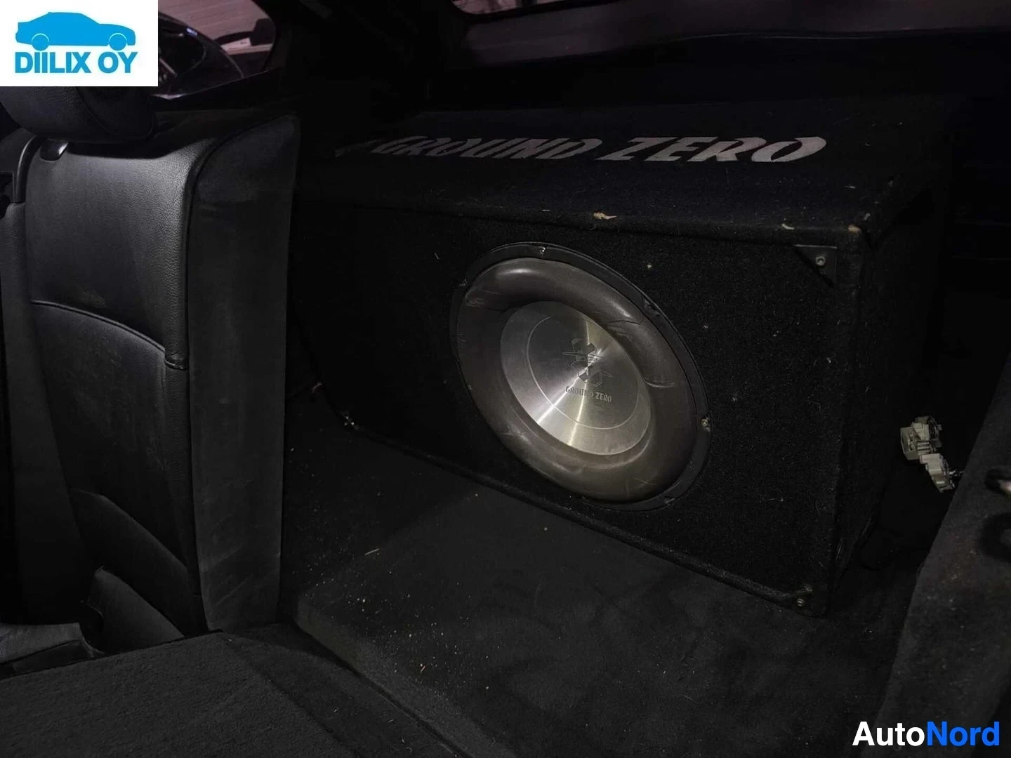 Ohjaamo: Kuva auton sisätilasta, jossa näkyy takapenkki ja tavaratilassa oleva iso subwoofer-kaiutin, jossa lukee "GROUND ZERO".
