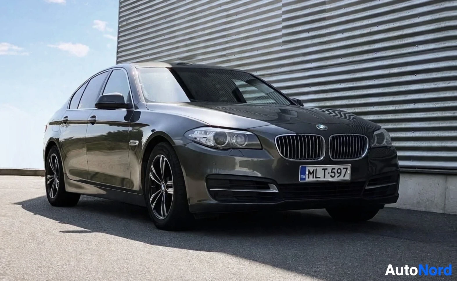 Etuviisto, nokka oikealle: Tummanharmaa BMW 5-sarjan sedan edestä vasemmalta, pysäköitynä rakennuksen eteen. Auton rekisterinumero on
