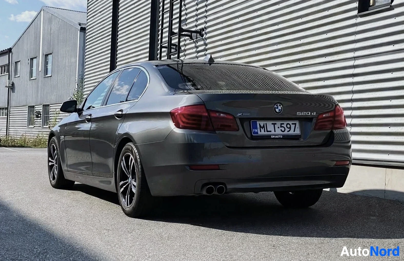 Takaviisto: Tummanharmaa BMW 5-sarjan sedan takaa vasemmalta, pysäköitynä rakennuksen eteen. Auton rekisterinumero on