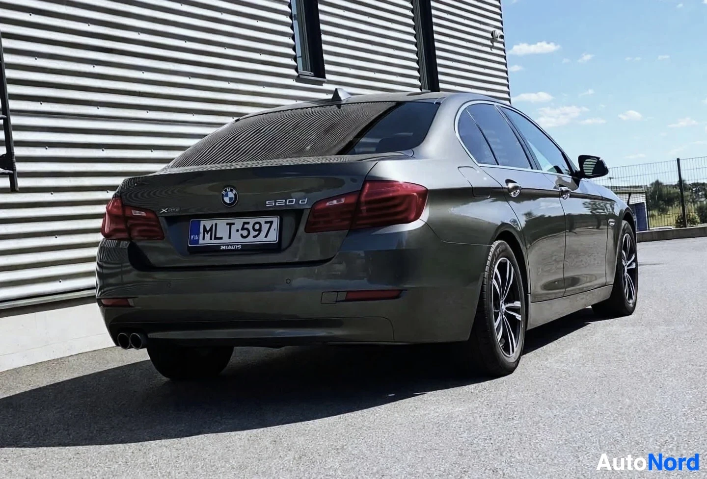 Takaviisto: Tummanharmaa BMW 5-sarjan sedan takaa oikealta, pysäköitynä rakennuksen eteen. Auton rekisterinumero on