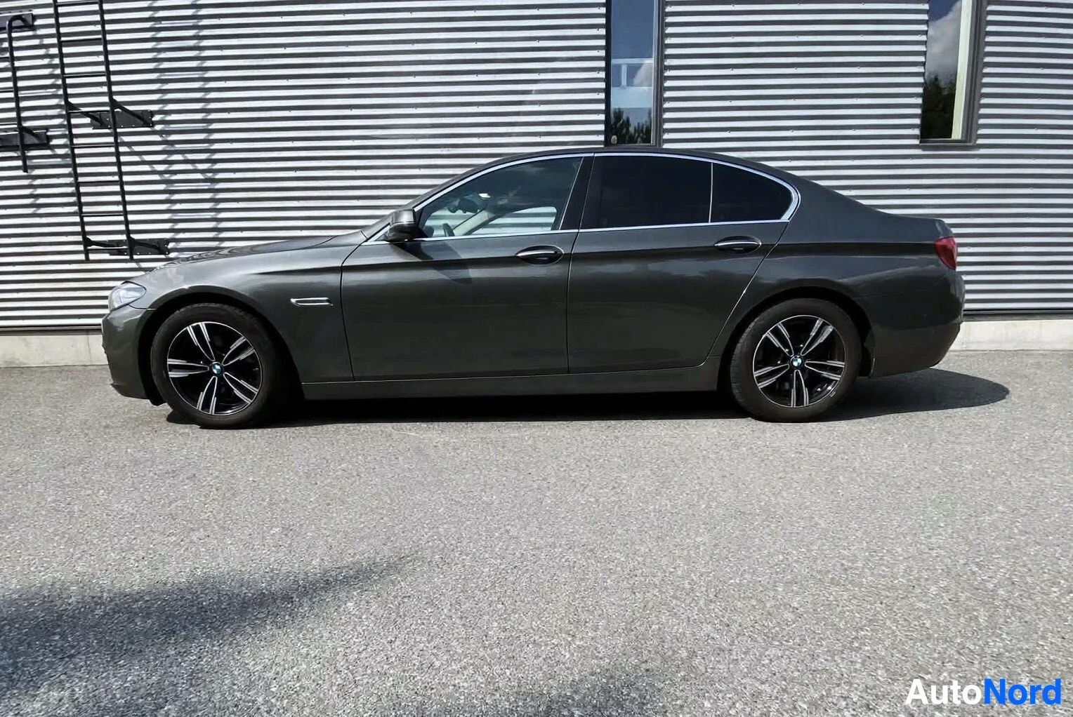 Sivulta: Sivukuva tummanharmaasta BMW 5-sarjan sedanista parkkipaikalla, jossa auton vasen kylki ja mustat monipuolaiset kevytmetallivanteet näkyvät.