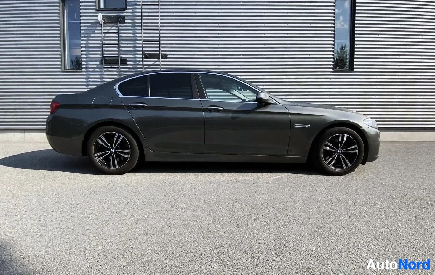 Sivulta: Sivukuva tummanharmaasta BMW 5-sarjan sedanista parkkipaikalla, jossa auton oikea kylki ja mustat monipuolaiset kevytmetallivanteet näkyvät.