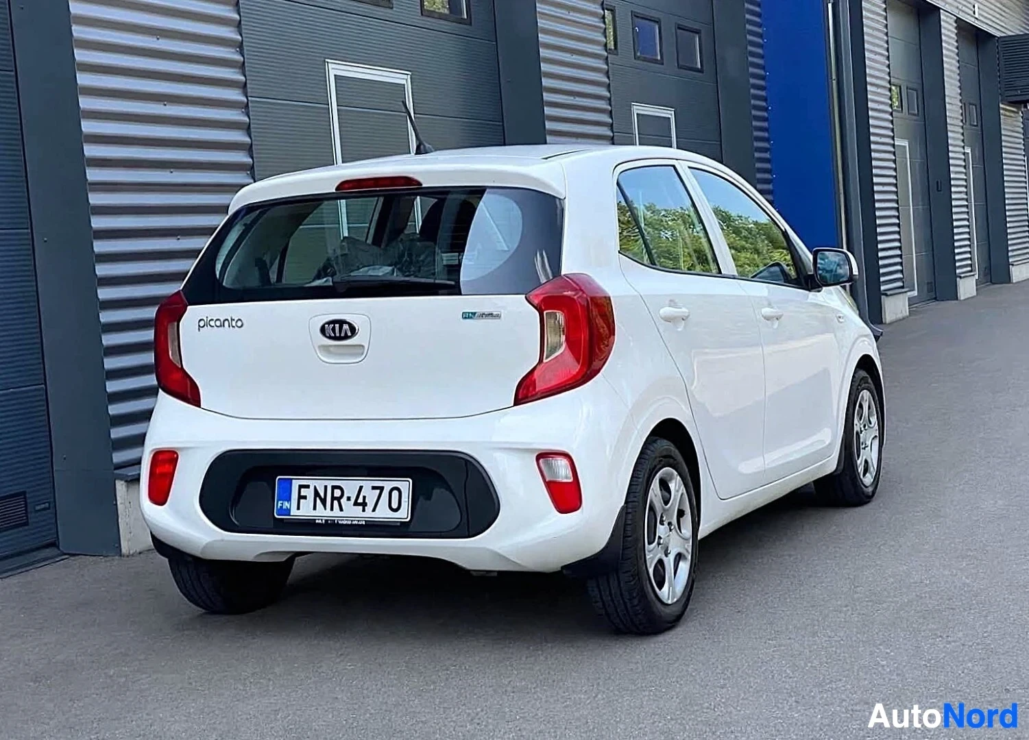 Takaviisto: Valkoinen Kia Picanto on pysäköity rakennuksen eteen, takaosa oikealle. Auton takaluukussa on "picanto"-merkki vasemmalla ja KIA-logo keskellä. Oikealla on "Eco