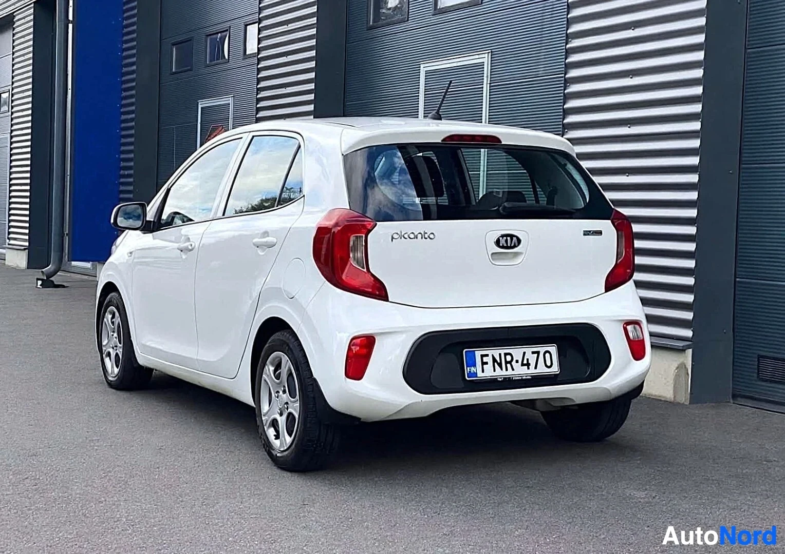 Takaviisto: Valkoinen Kia Picanto on pysäköity rakennuksen eteen, takaosa oikealle. Auton takaluukussa on "picanto"-merkki vasemmalla ja KIA-logo keskellä. Rekisterikilpi F