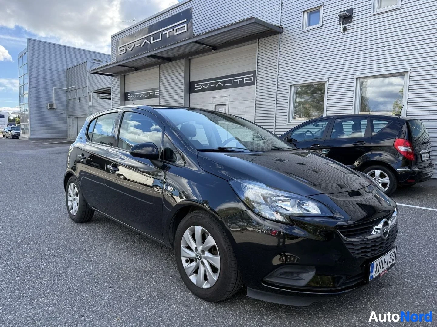 Etuviisto, nokka vasemmalle: Musta Opel Corsa -henkilöauto näkymä etukolmesta vasemmalta, paikoitusalueella SV-AUTO-huoltoliikkeen edessä