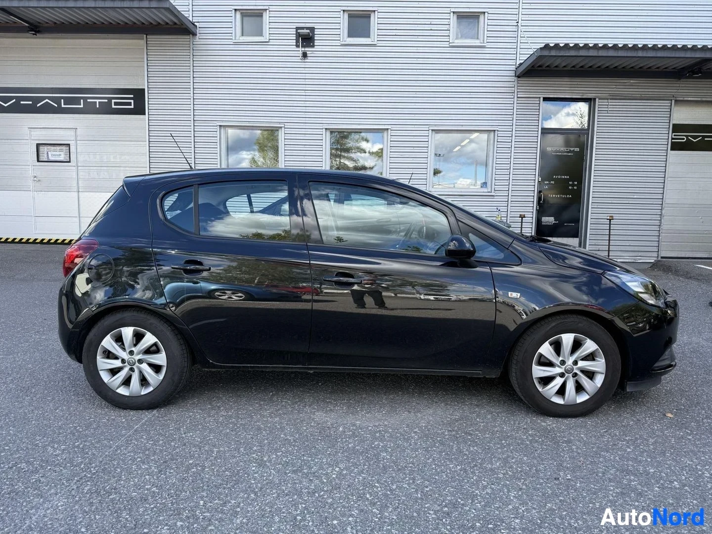 Sivulta: Musta viisisuinen henkilöauto sivusta katsottuna, valkoilla vantein, pysäköitynä graanitilalla. Taustalla teollisuusrakennuksia.