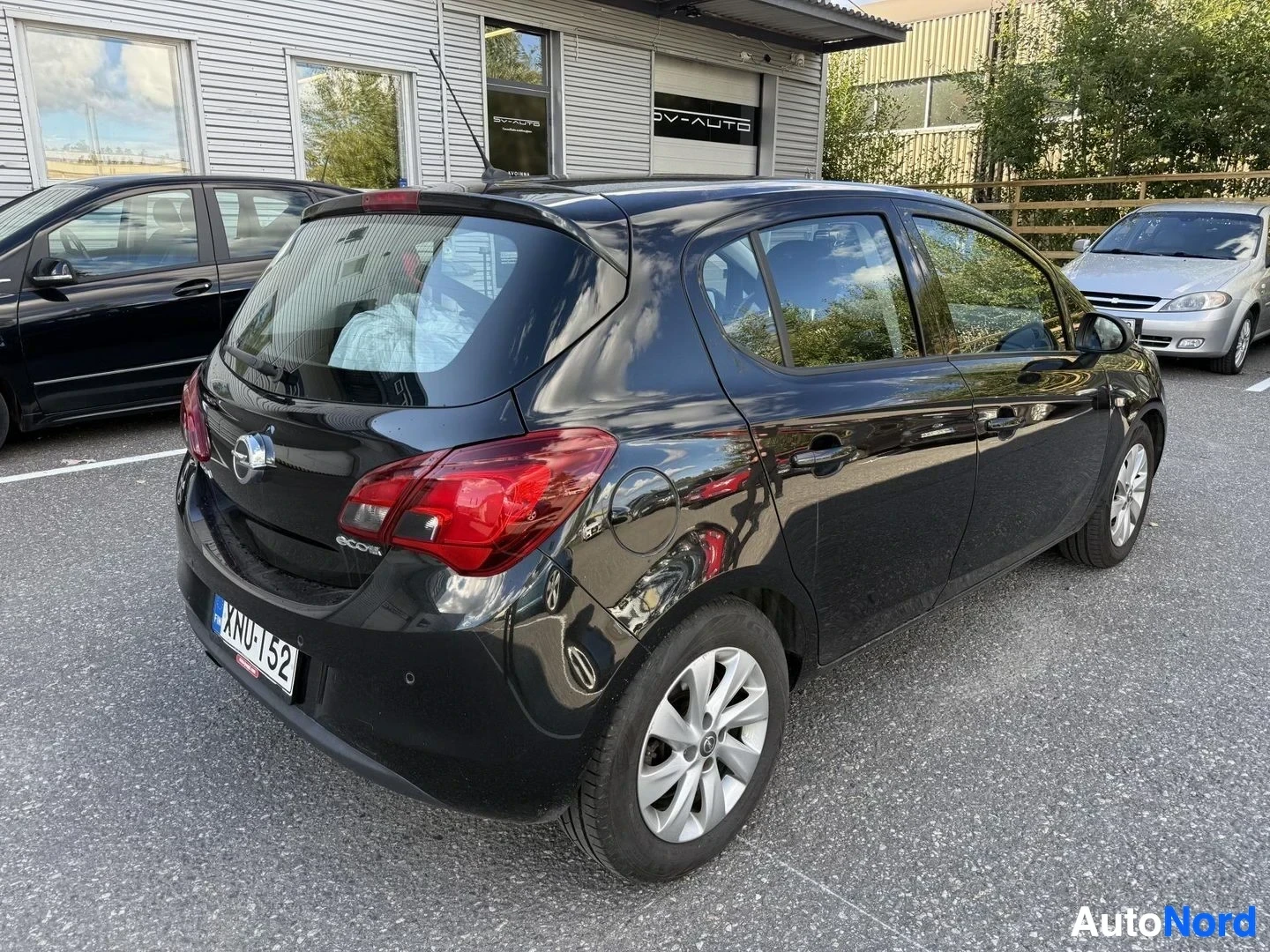 Takaviisto: Musta pieni katuauto näkyy oikealta takakolmionäkymältä; näkyviin pääsee takaovi, takalasin ja osa sivustasta. Tummansinisten vanteiden valkoinen pinta ja punai