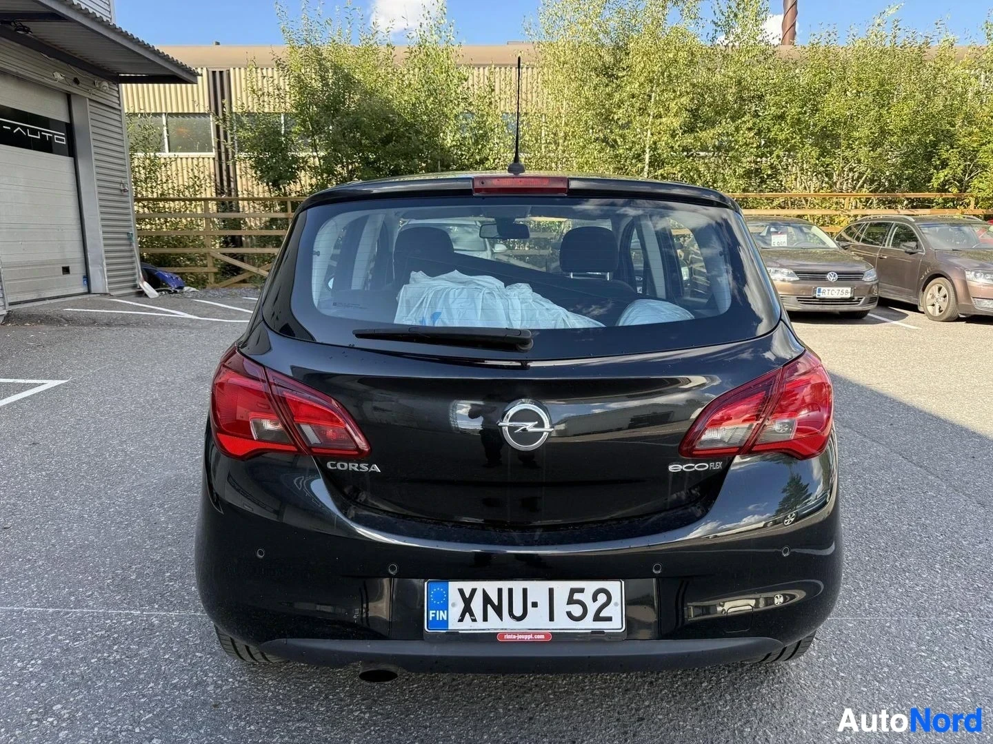 Takaa: Musta Opel Corsa takaa kuvattuna, rekisterikilpi näkyvillä, punaiset takavalot, pysäköintialue taustalla
