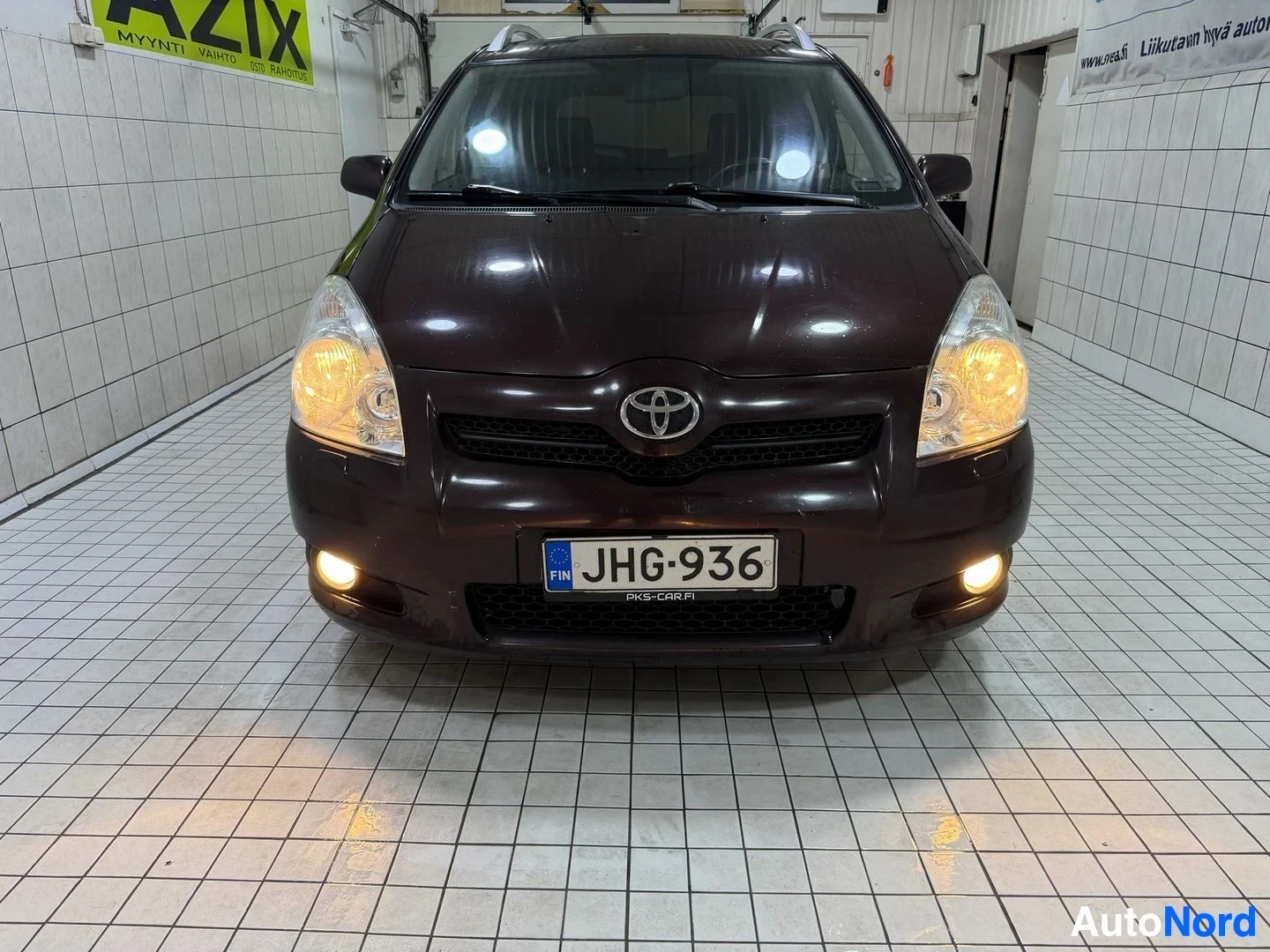 Edestä: Tummanruskea Toyota-auto, etupuoli, sisätiloissa valkoisen laatoituksen edessä. Rekisterinumero näkyy. Kaukovalojen valot syttyneinä