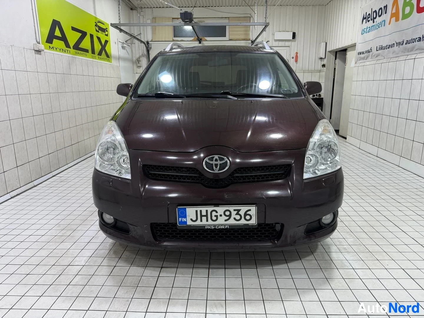 Edestä: Toyota-auto edestä katsottuna sisätiloissa, tummanruskea väri, valkoset ajovalot, rekisterikilpi