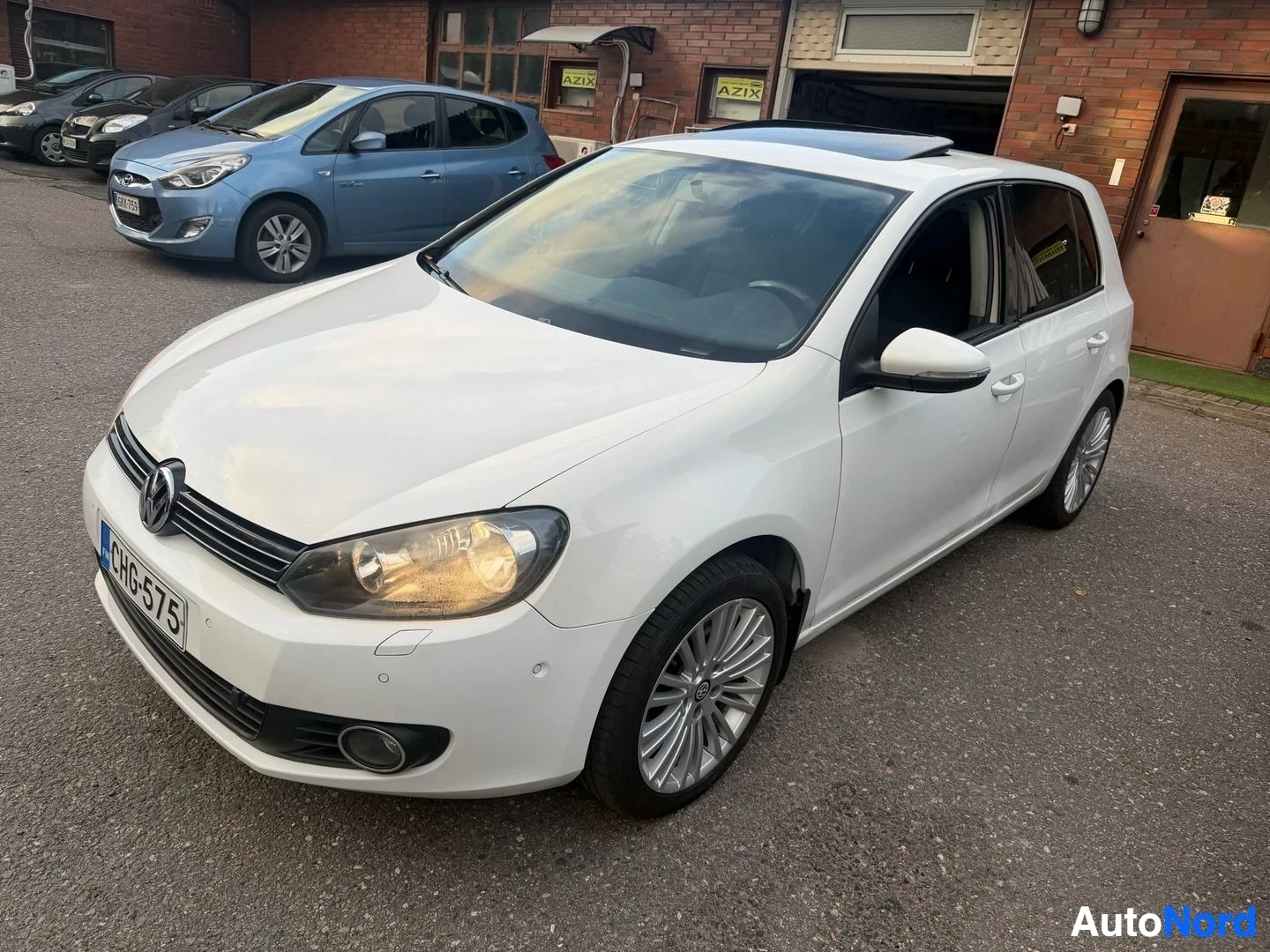 Etuviisto, nokka vasemmalle: Valkoinen Volkswagen Golf -pienoisperhesauto pysäköitynä pihalla, kuvattuna edestä vasemmalta puolelta. Näkyvissä etupää, kattokupla ja oikea sivu. Rek