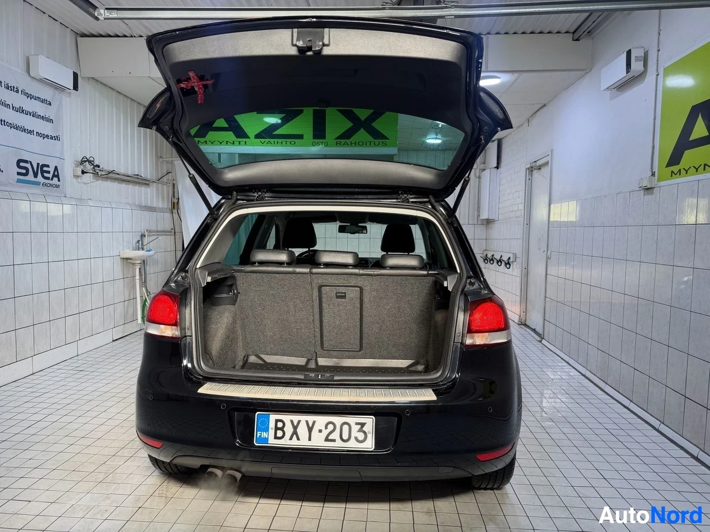 Tavaratila: Musta Volkswagen Golf-tyyppinen viisipaikkainen auto, takaluukku auki, osoittaa lastauatilaa ja rekisteritunnusta