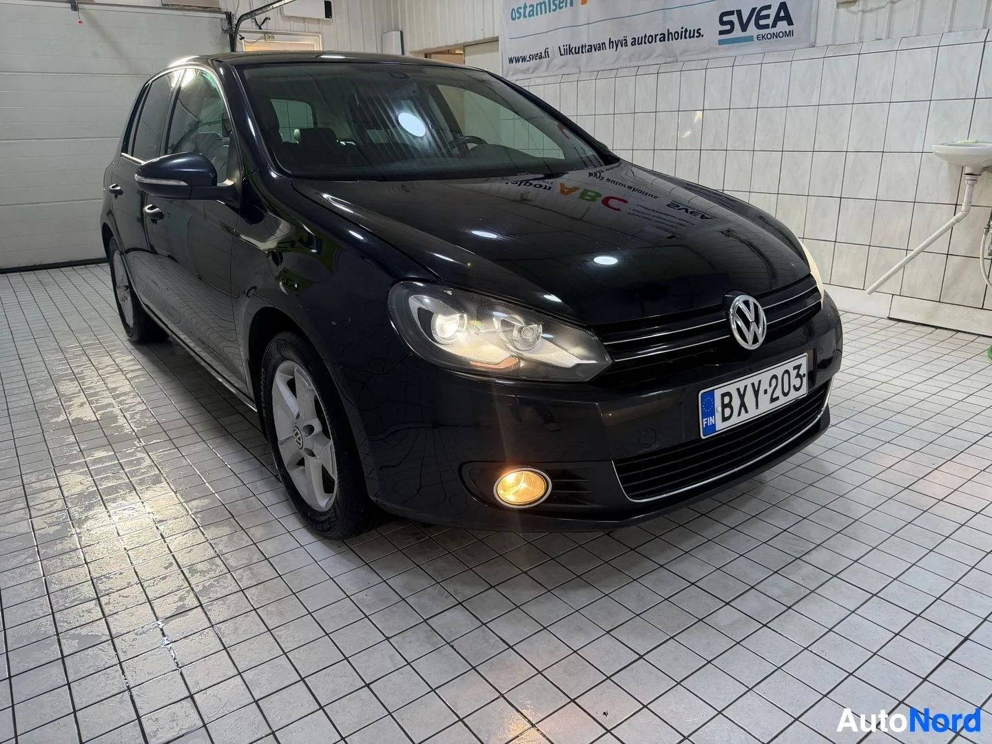 Etuviisto, nokka oikealle: Musta Volkswagen Golf kolmen neljäsosan näkymä vasemmalta puolelta. Rekisterikilvessä. Ajovalot palavat. Kattava näkymä koko ajoneuvosta varikon