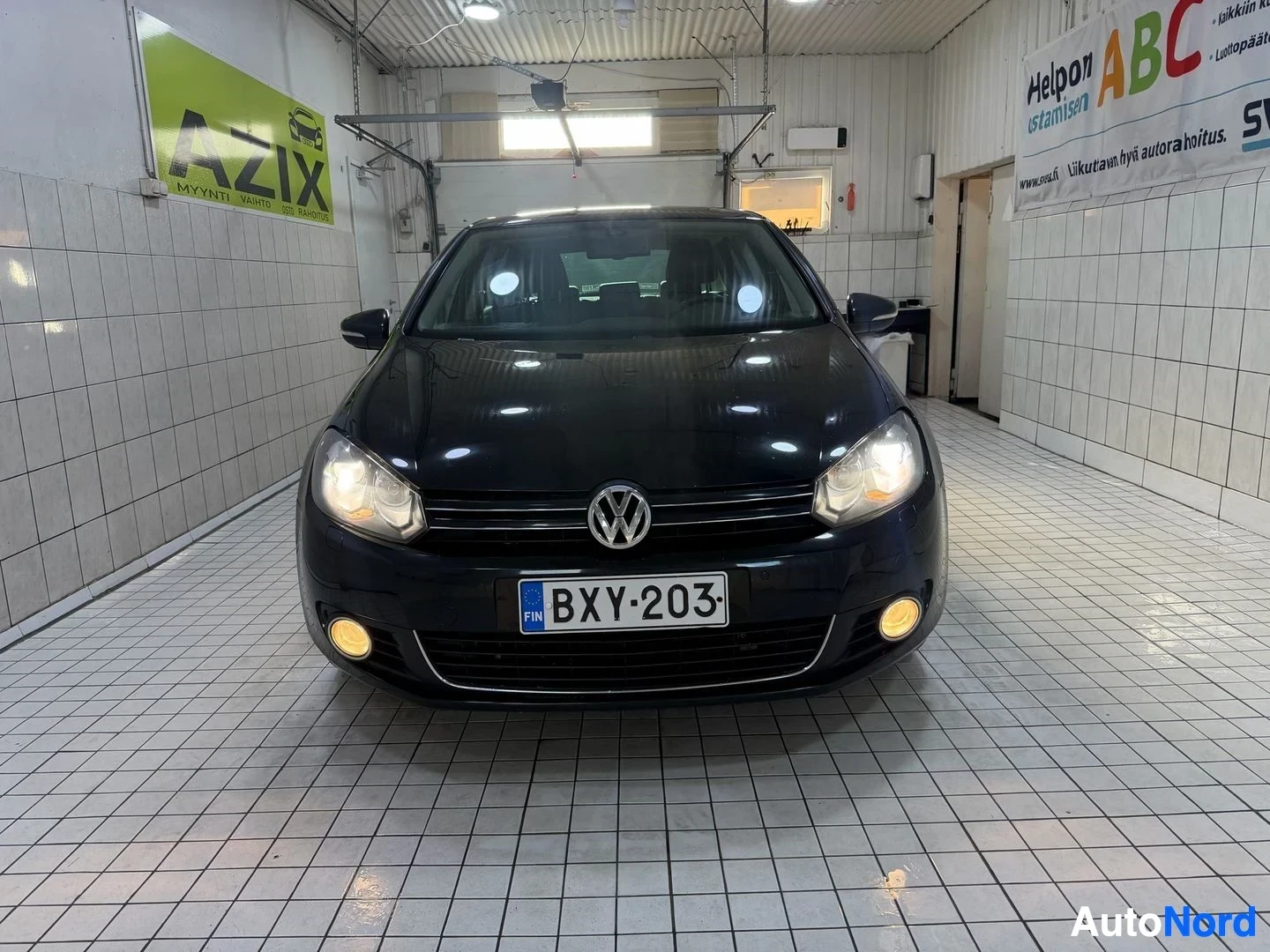 Edestä: Musta Volkswagen Golf edestä katsottuna varikon sisällä. Rekisterikilit näkyvät. Ajovalot palavat. Koko ajoneuvo näkyvä