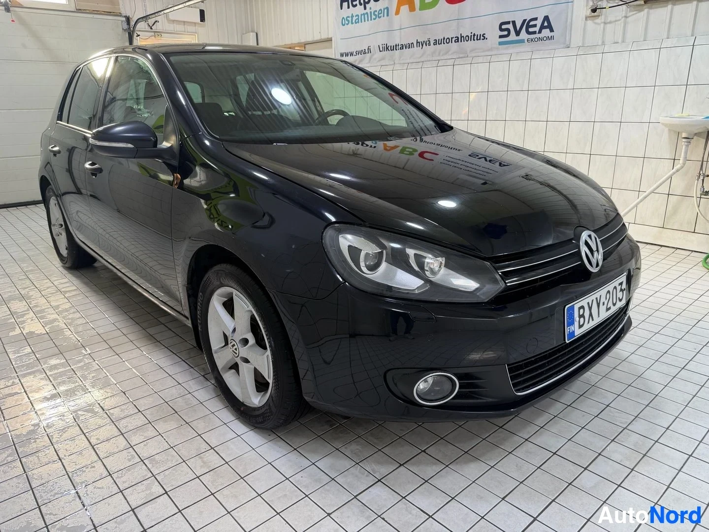 Etuviisto, nokka vasemmalle: Musta Volkswagen Golf etulta kolmen neljänneksen kulmasta vasemmalle osoittaen, sisätiloissa. Rekisterikilpi näkyvissä