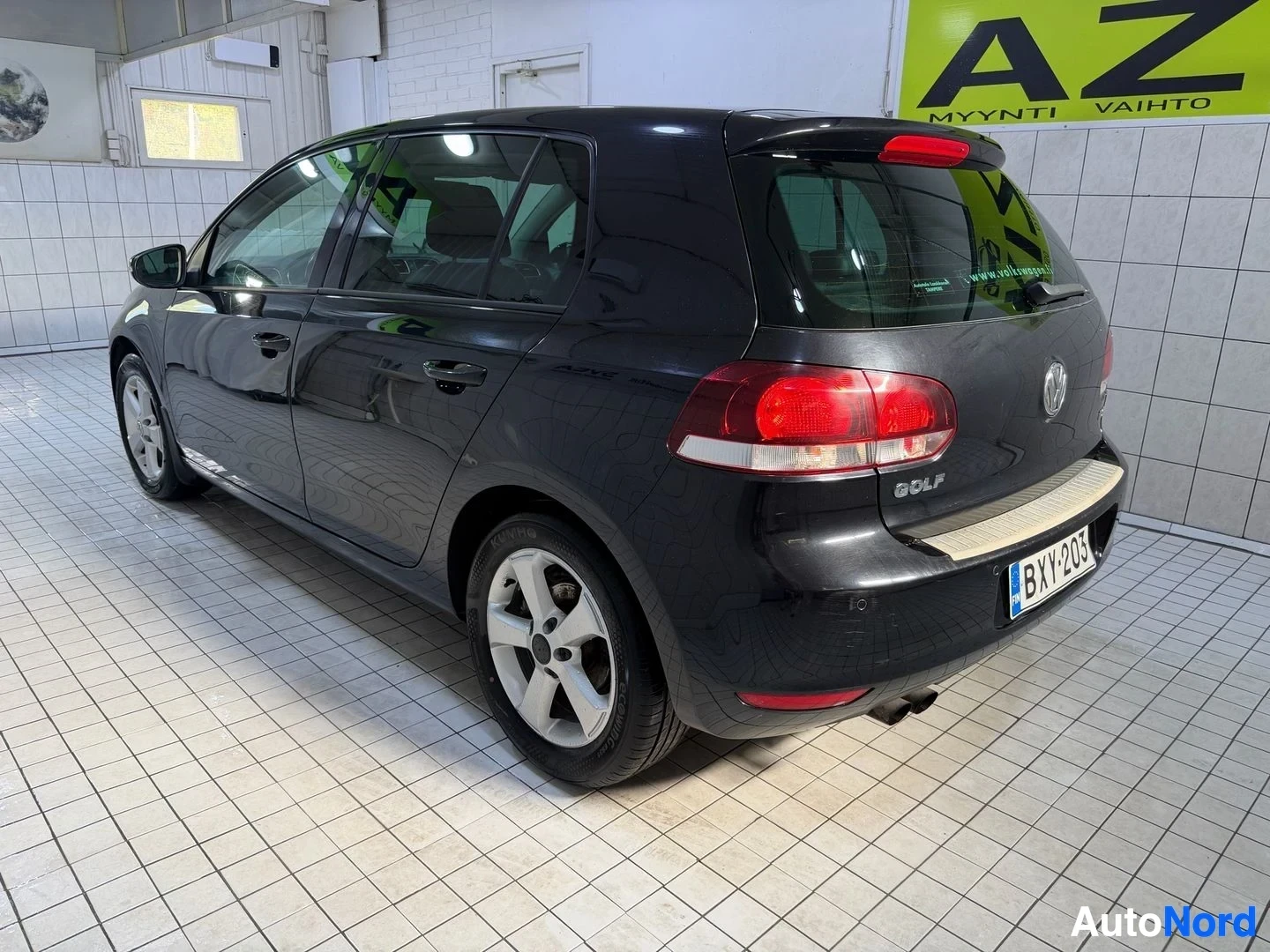 Takaviisto: Musta Volkswagen Golf takaa kolmen neljänneksen kulmasta oikealle osoittaen, sisätiloissa. Ovi avoin. Rekisterikilpi näkyvissä