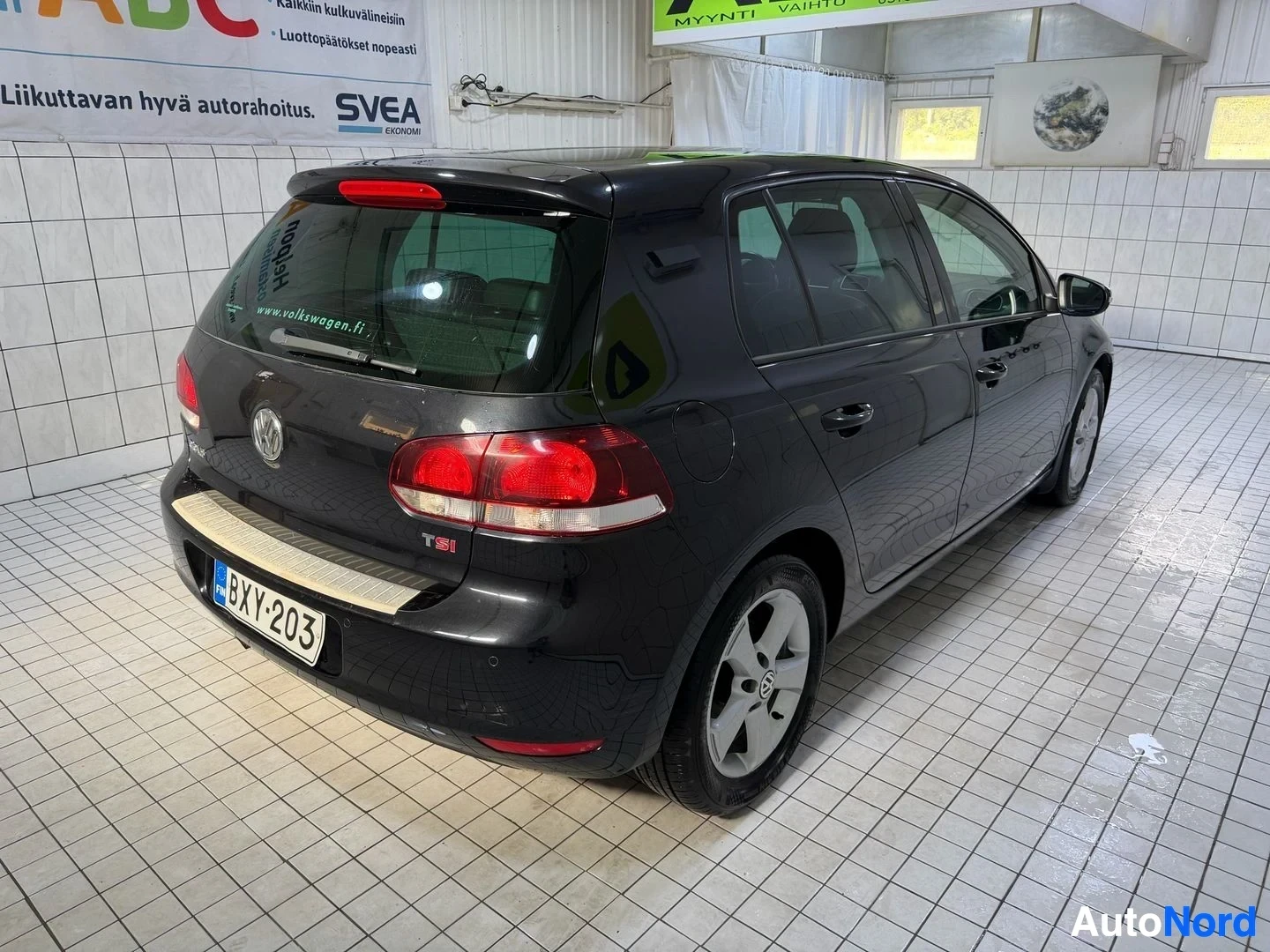 Takaviisto: Musta Volkswagen Golf takaa kolmen neljänneksen kulmasta vasemmalle osoittaen, sisätiloissa. Rekisterikilpi näkyvissä