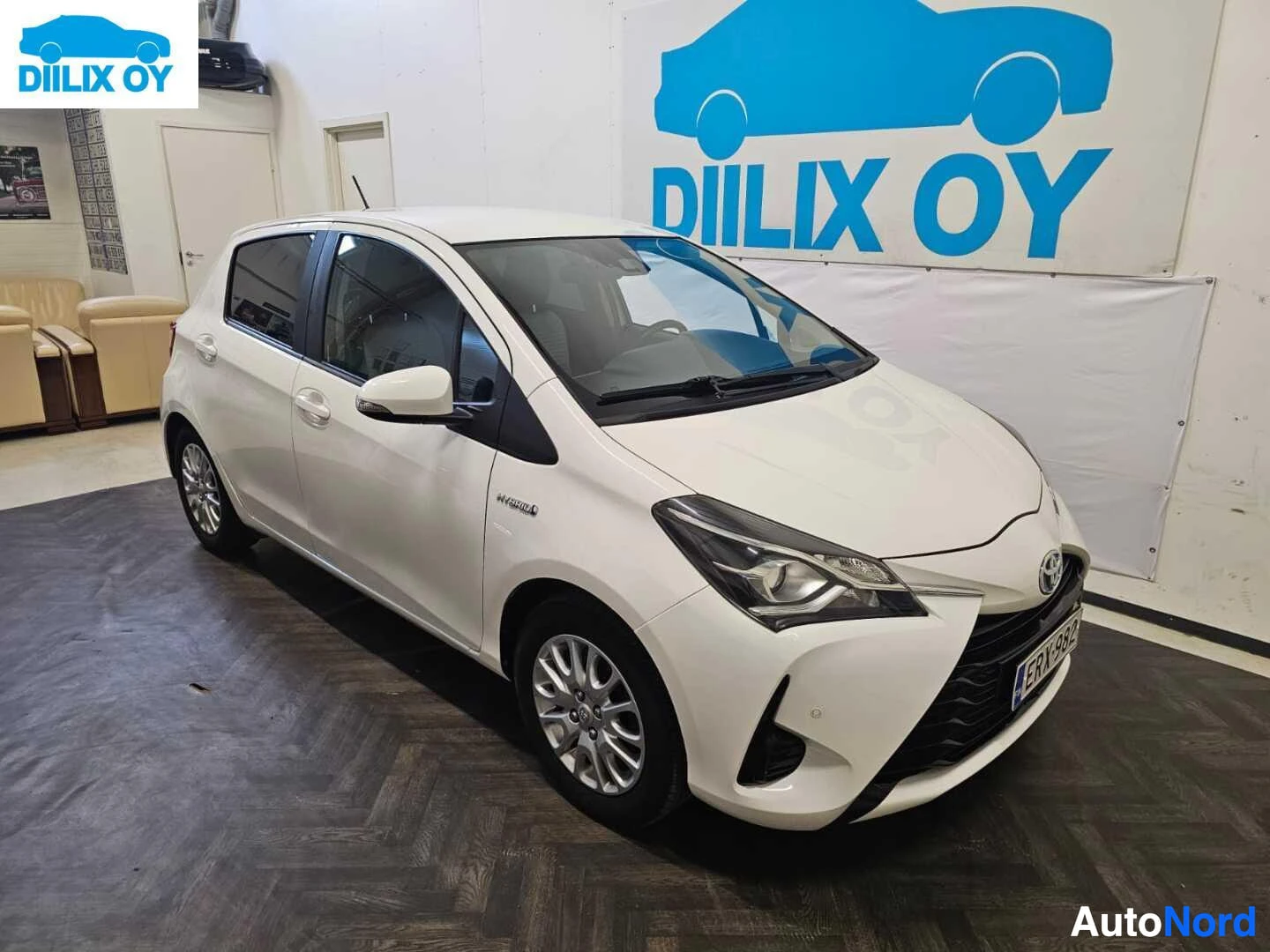 Etuviisto, nokka oikealle: Valkoinen Toyota Yaris viistosti edestä oikealta kuvattuna sisätiloissa.