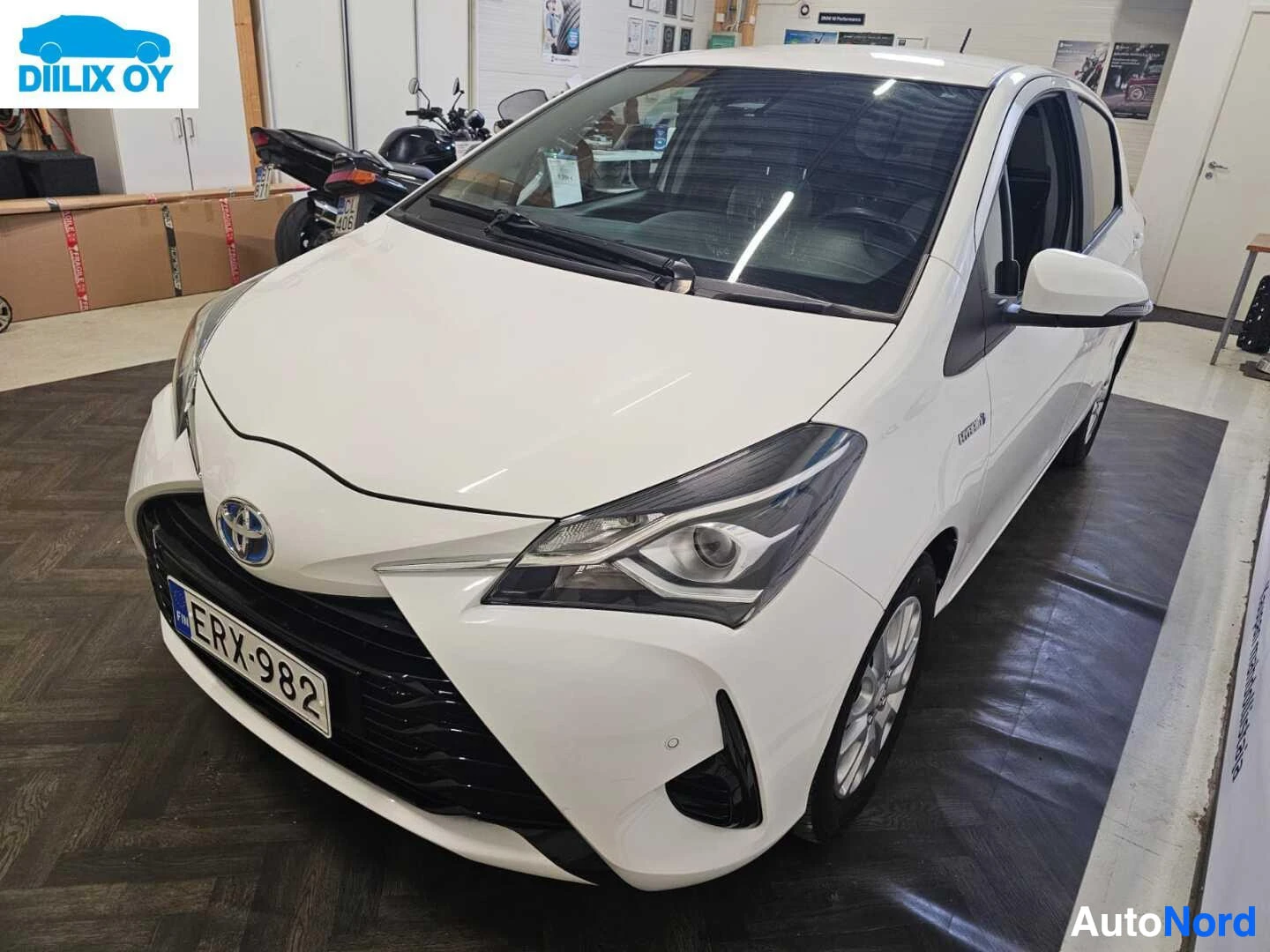 Etuviisto, nokka vasemmalle: Valkoinen Toyota Yaris, etupuoli vasemmalta kuvattuna. Rekisterikilpi näkyy. Auto on näyttelytilassa