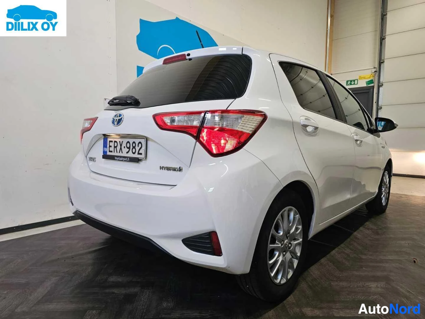 Takaviisto: Valkoinen Toyota Yaris takaa kuvattuna. Rekisterikilpi näkyy. Takavalot punaisina. Auto on hallissa