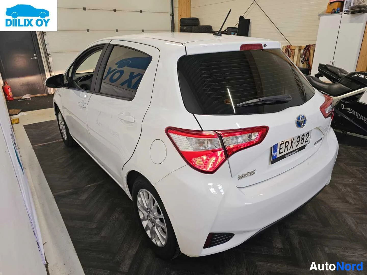 Sivulta: Valkoinen Toyota Yaris oikealta sivulta kuvattuna. Rekisterikilpi näkyy. Auto on hallissa, ovi on auki