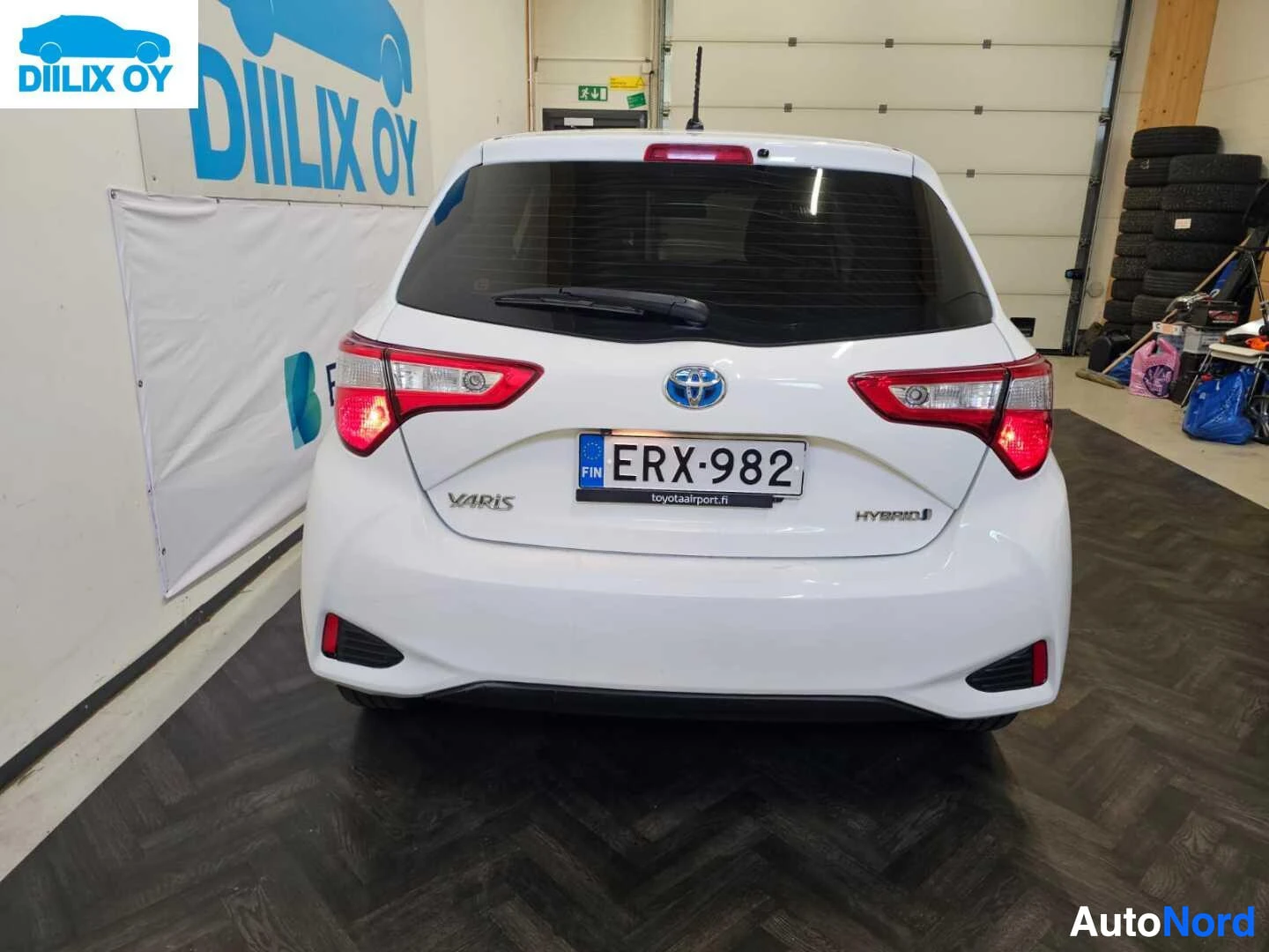 Takaa: Valkoinen Toyota Yaris Hybrid, takapuoli hallissa, rekisteri näkyy, punaisten takavalojen ja hybrid-merkinnän kanssa