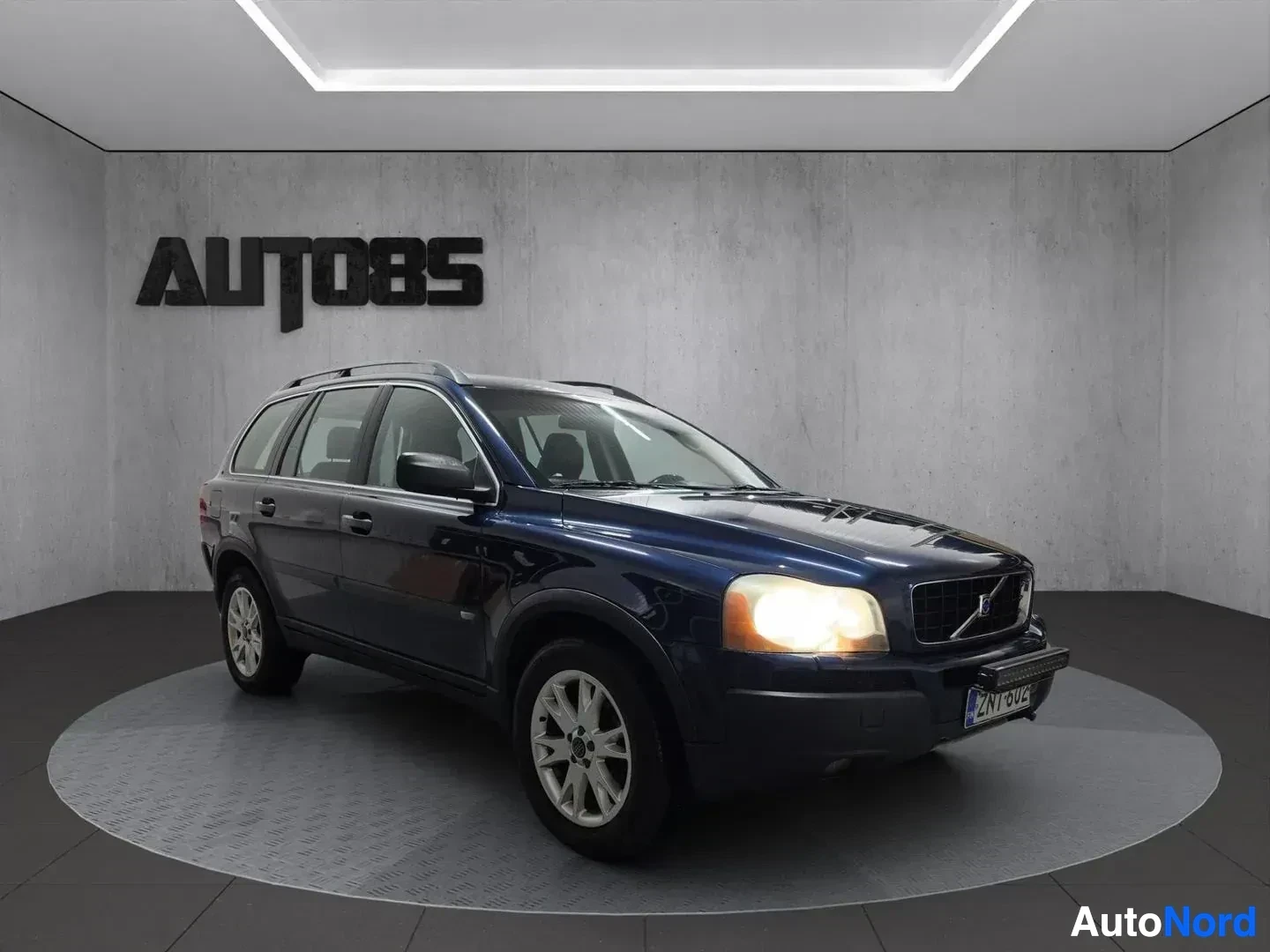 Etuviisto, nokka oikealle: Siniset Volvo XC90:n etu- ja sivukulma, ajovalot päällä, valaistussa studiossa. Auton katolla on kattotelineet.
