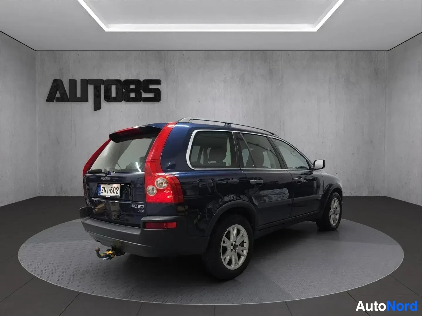 Takaviisto: Sivunäkymä sinisestä Volvo XC90 -autosta, jossa on vetokoukku, auton perä vasemmalla ja keula oikealla. Auto on kuvattu harmaata seinää vasten valaistussa huone