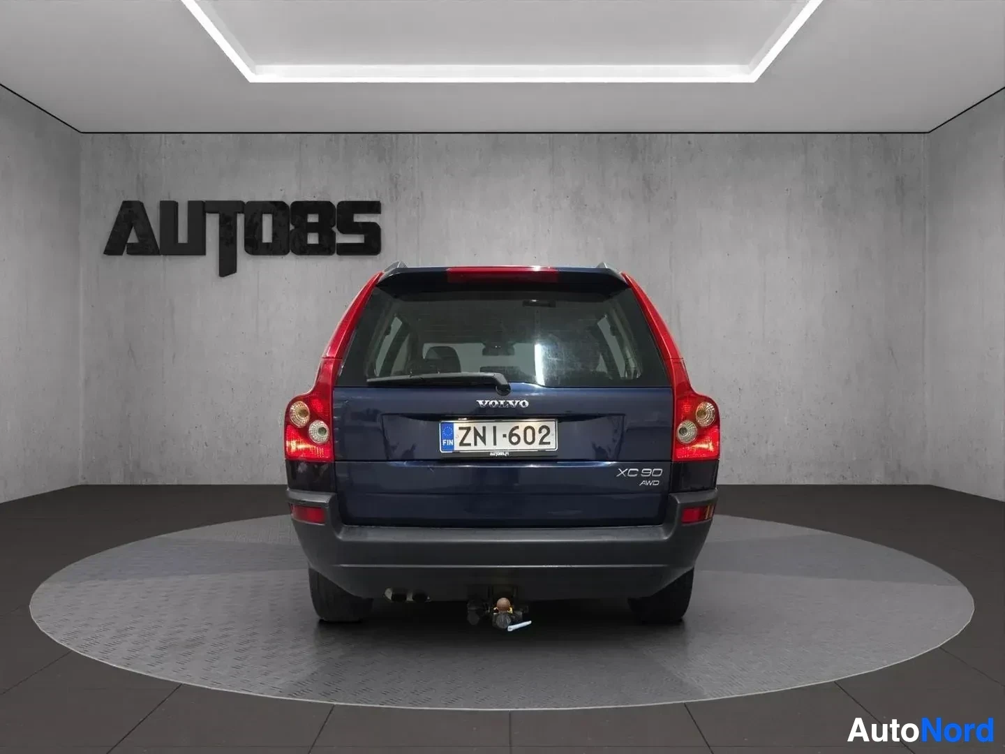 Takaa: Volvo XC90 -auton takaosa, jossa on vetokoukku ja rekisterikilpi. Auto on kuvattu harmaata seinää vasten valaistussa huoneessa