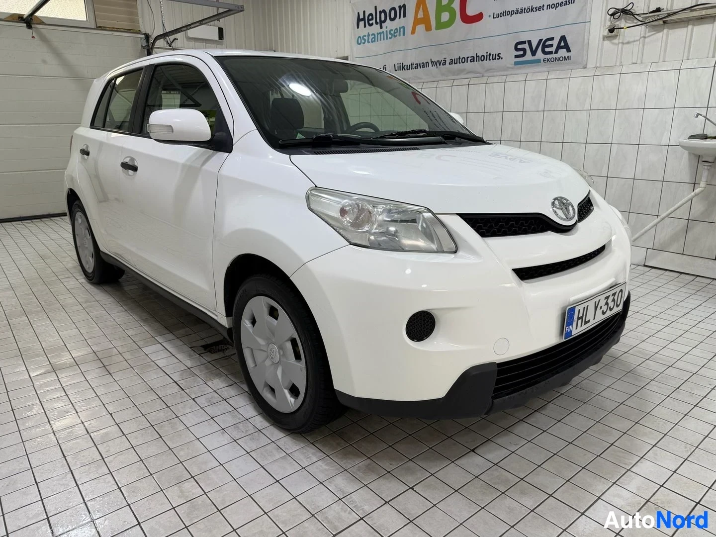 Etuviisto, nokka vasemmalle: Valkoinen Toyota Urban Cruiser, etupuoli vasemmalta, pysäköity sisäkattoisessa hallissa, näkyy ABC-merkin yllä.