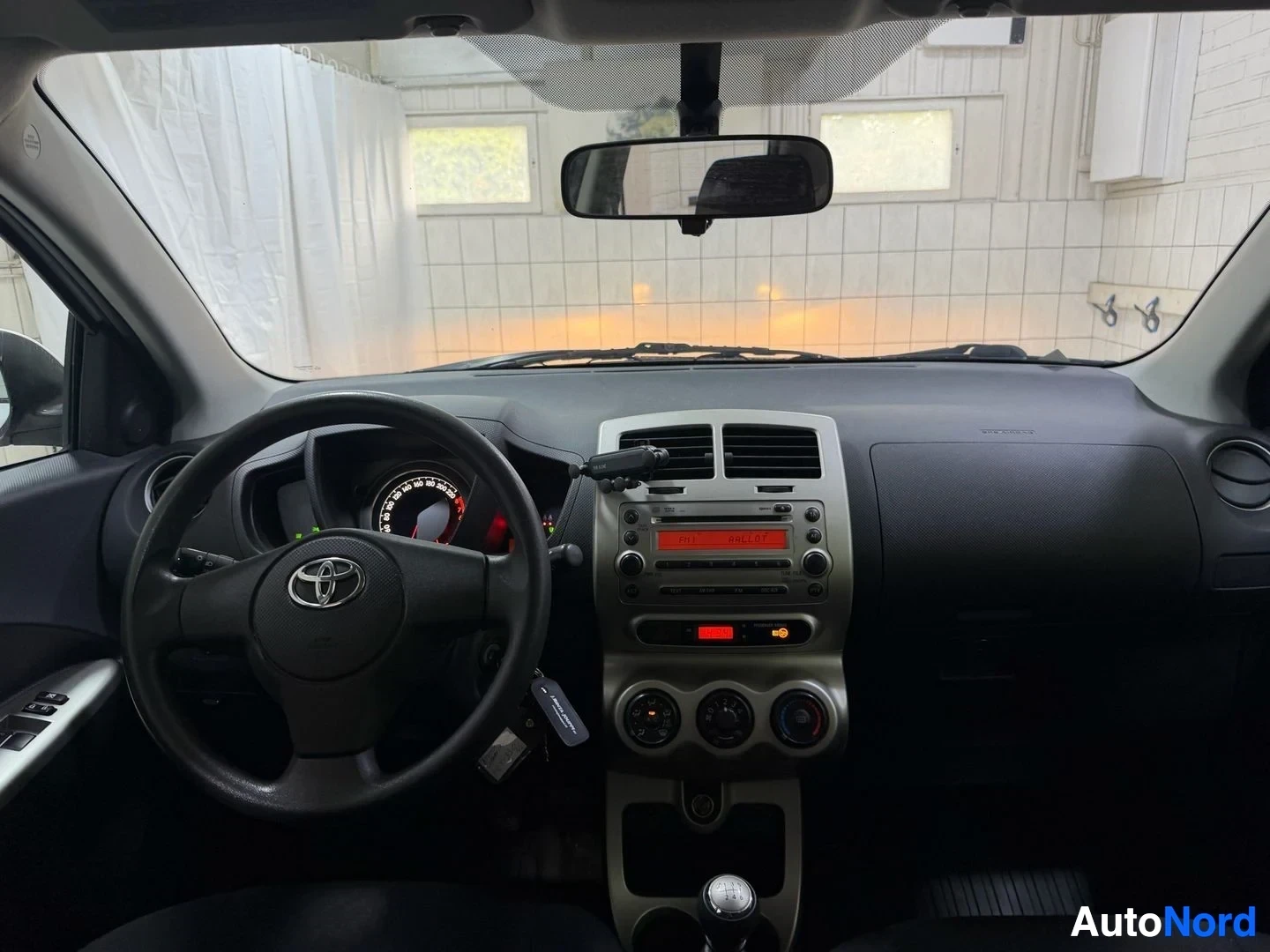 Ohjaamo: Auton sisätila etupuolelta: Toyota-merkki ohjauspyörässä, harmaa kojelauta, vaaleansävyinen infotainment-yksikkö punaisella näytöllä, kolmen kierteen ilmastoint