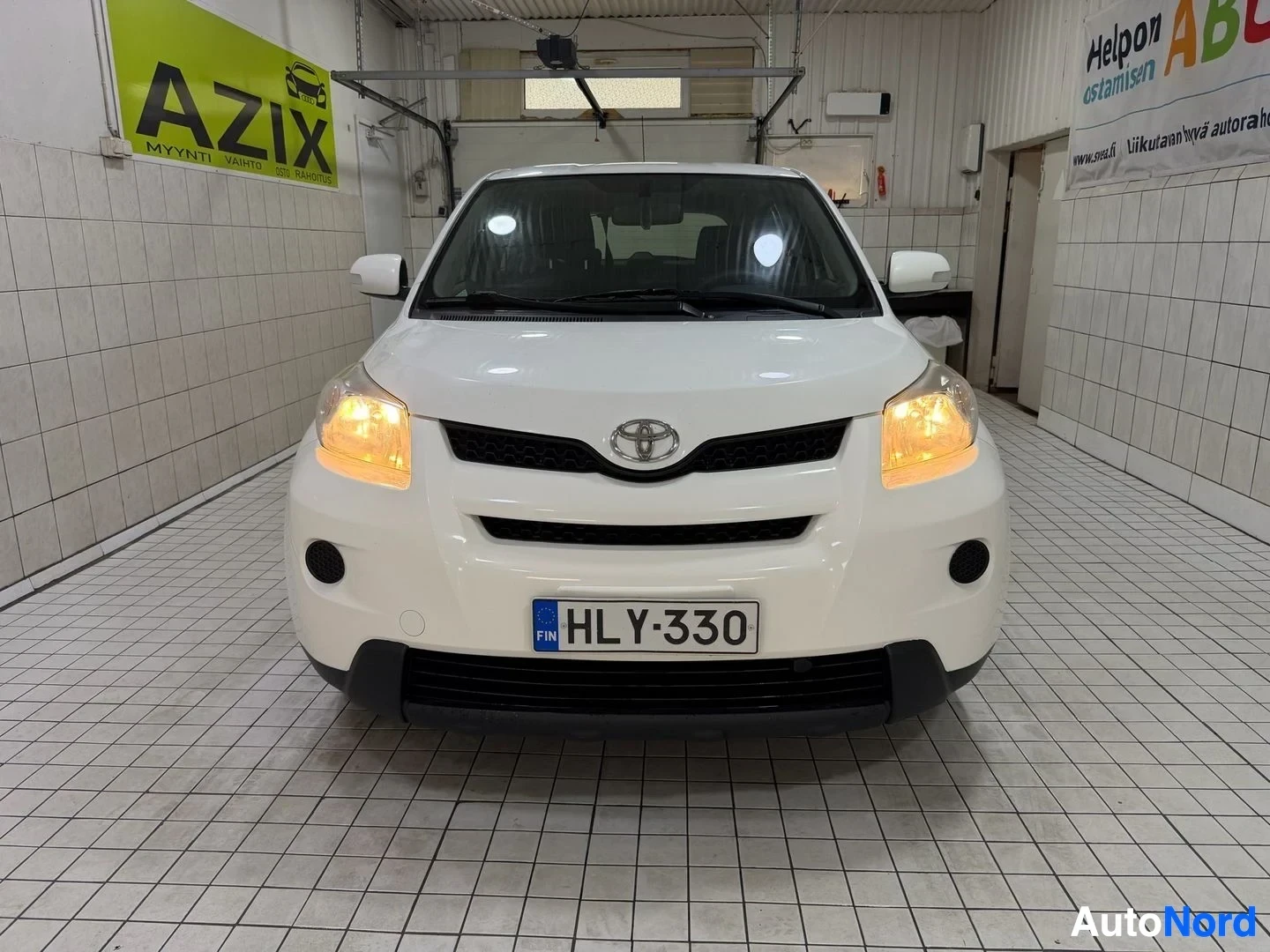 Edestä: Valkoinen Toyota-autolamolla kylpi varikolla. Etunäkymä, auto näkyy kokonaan. Rekisterikilpi näkyvissä