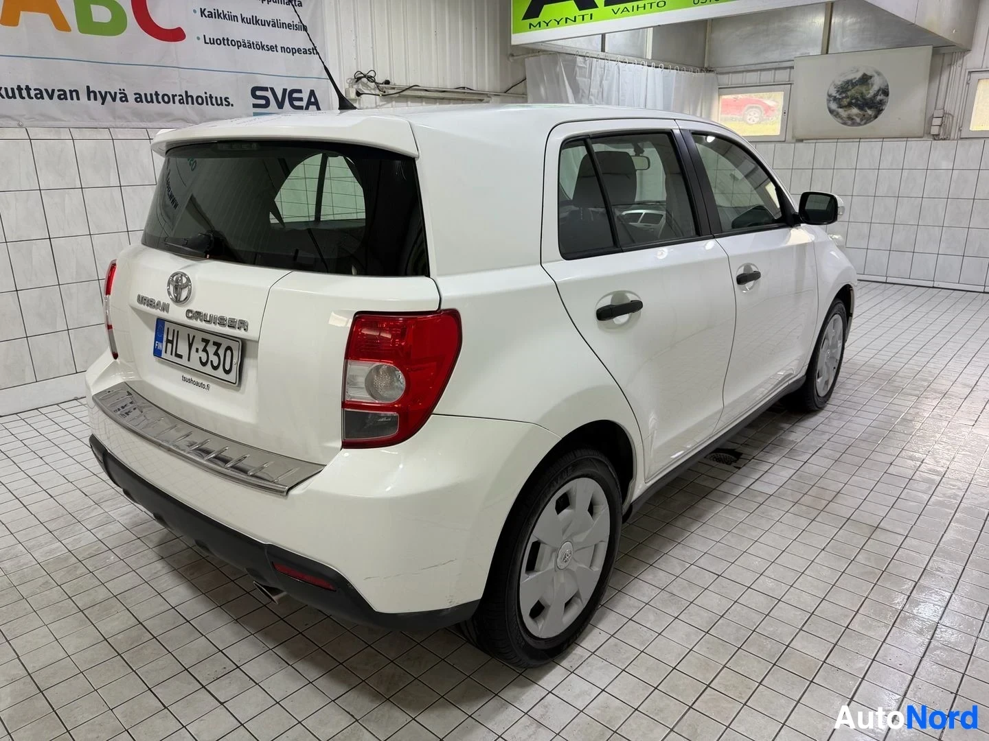 Takaviisto: Valkoinen Toyota Urban Cruiser, takapuoli oikealta, näkyy koko auton sivu ja takaosa hallissa.