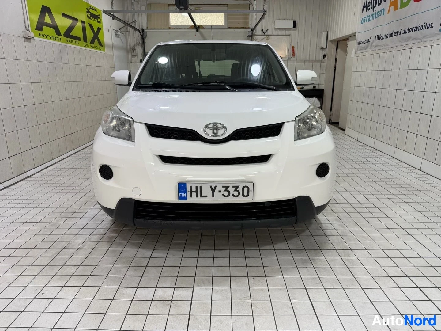 Edestä: Valkoinen pieni nelivetoiset auto, suora etunäkymä hallissa. Rekisterikilvessä lukee. Toyota-logo näkyy keskellä ajoneuvon etusillaa