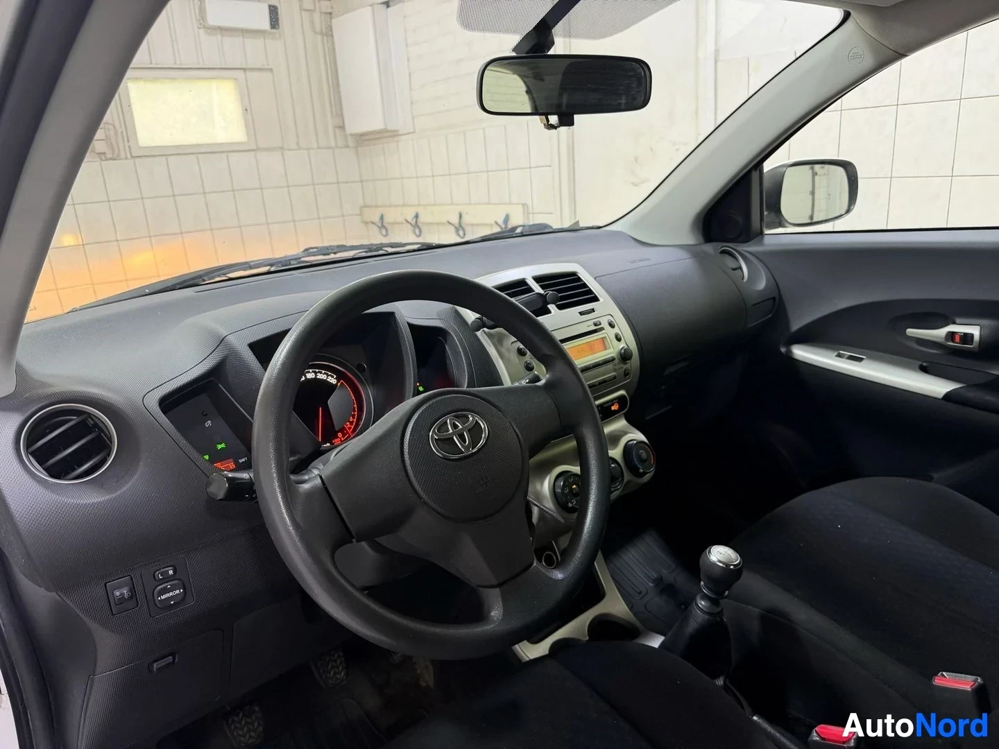 Ohjaamo: Auton sisätila etupuolelta: Toyota-merkki ohjauspyörässä, instrumentiklusterissa punainen neulanäyttö, keskikonsolissa vaaleansävyinen infotainment-yksikkö, mus